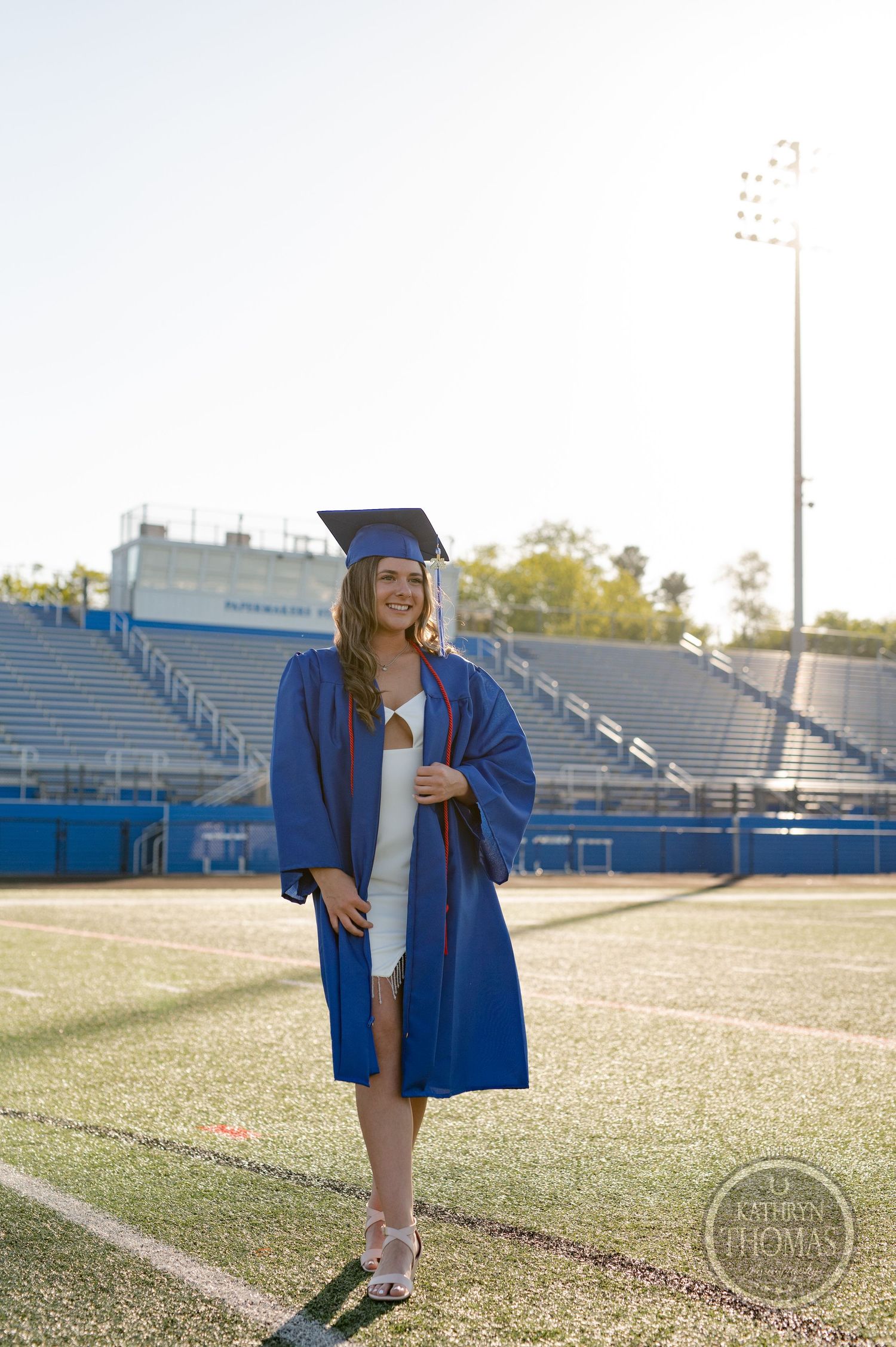 Graduate in blue gown poses on football field with stadium bleachers in background.