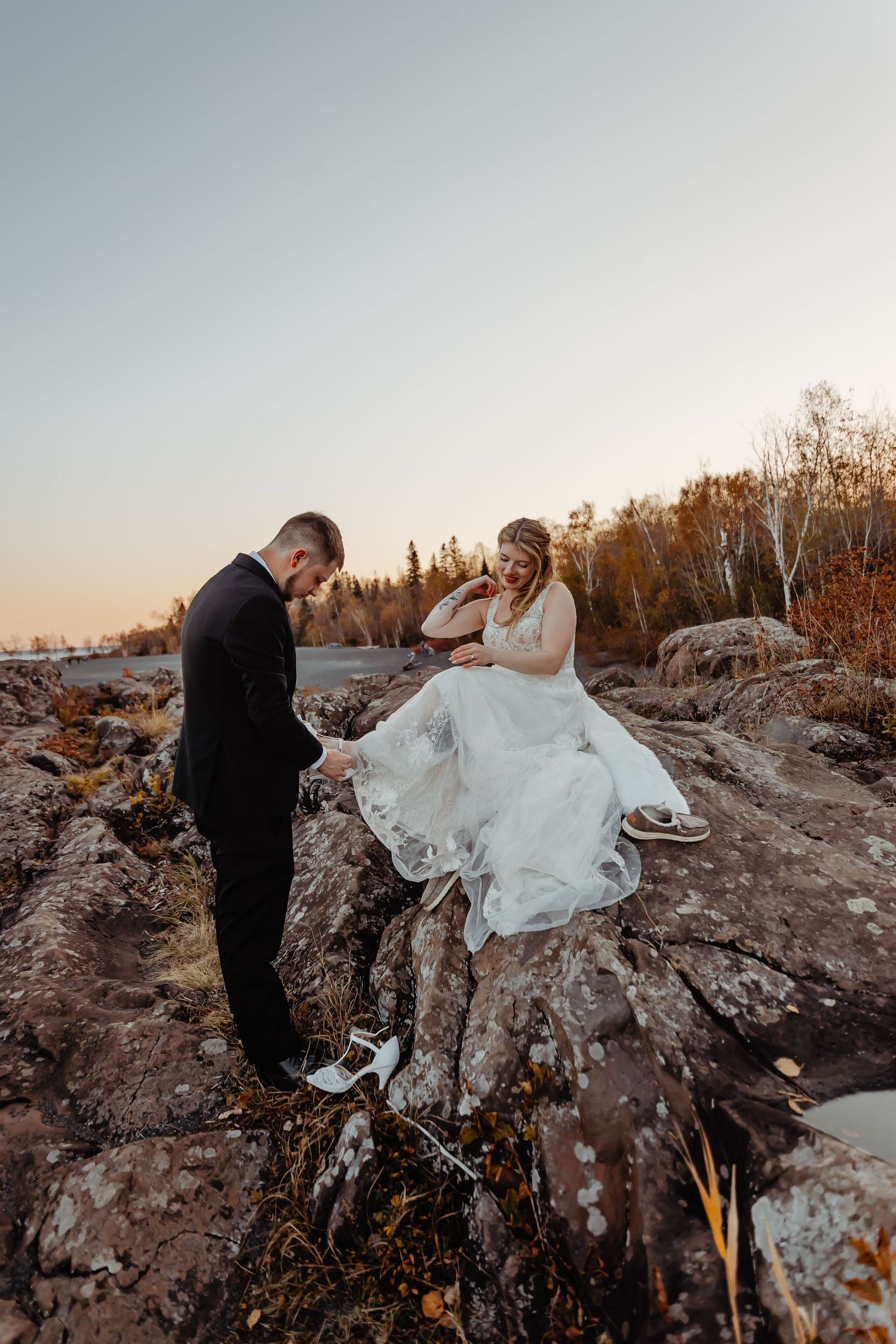 Couple in wedding attire standing on rocky shore during golden hour.