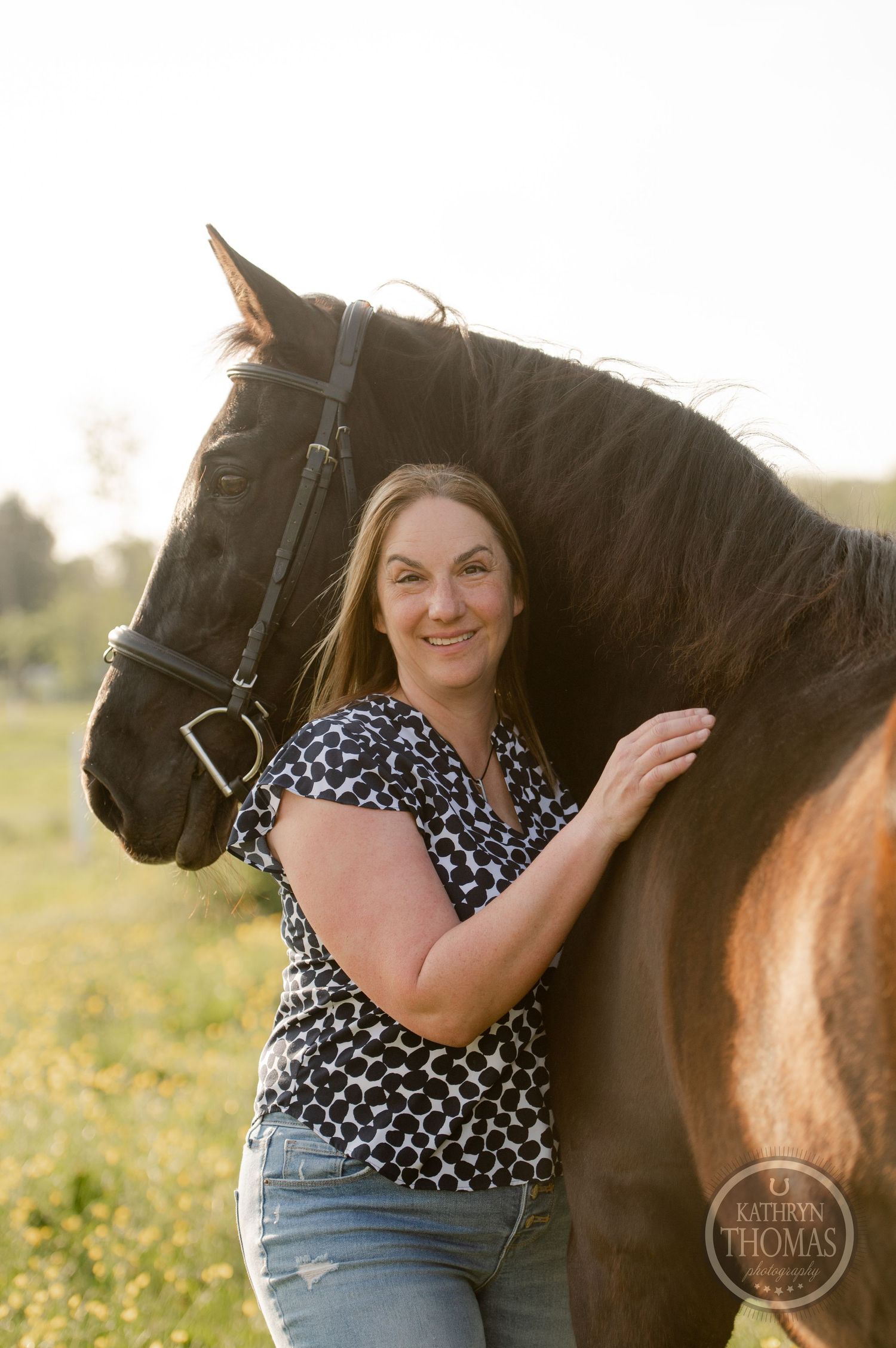 A beautiful dark horse stands with its caretaker in a floral top against a pastoral backdrop.