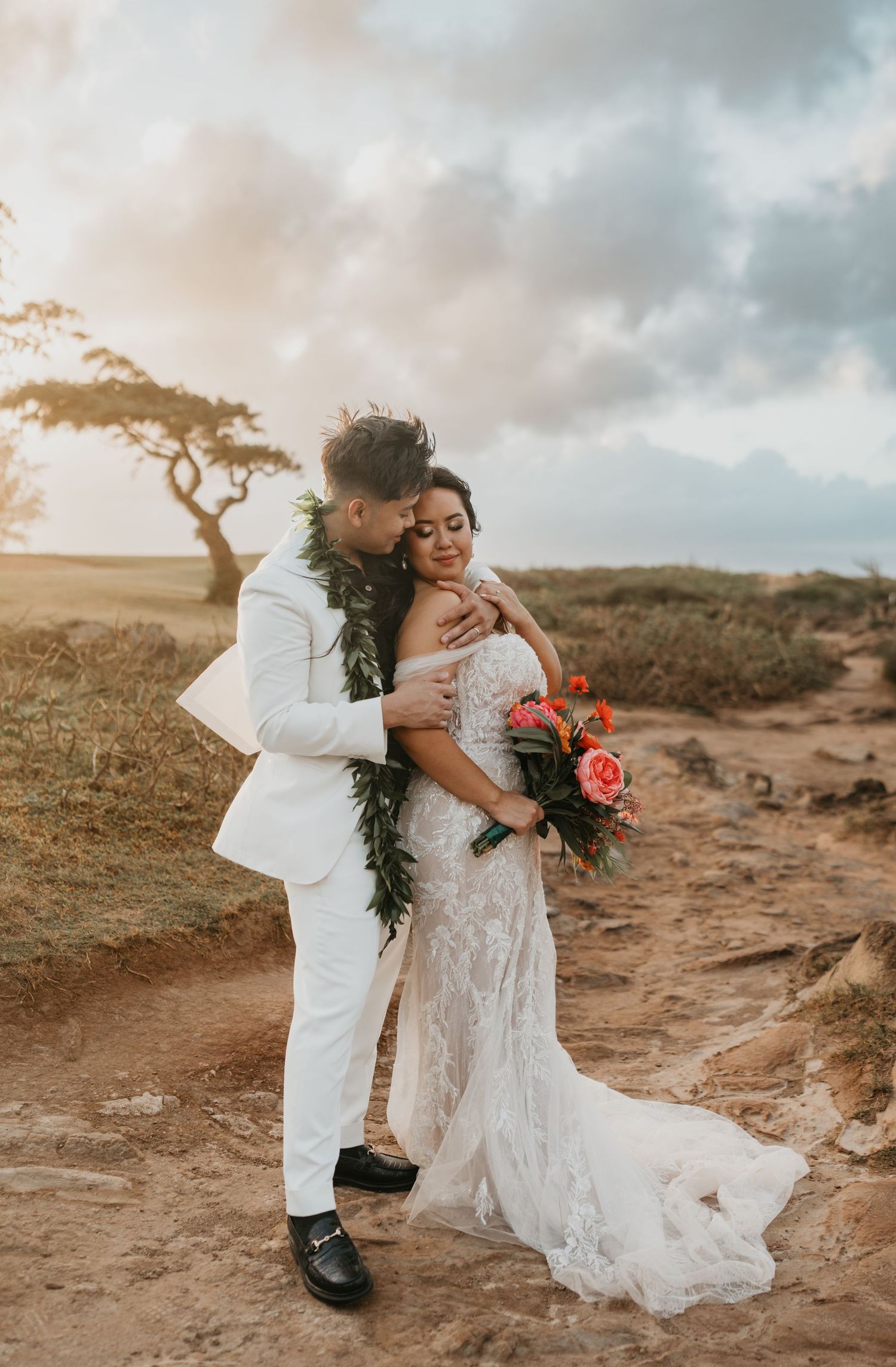 A romantic sunset wedding photo with a couple embracing on a cliff in Hawaii with a lone tree and orange sky.