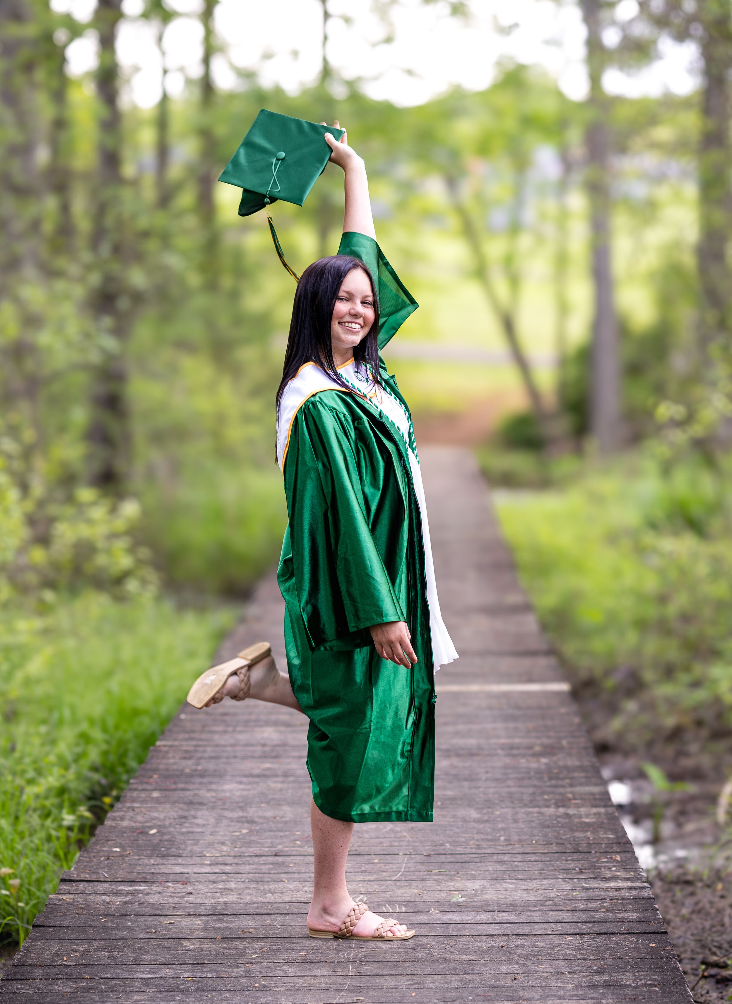 Graduate in green cap and gown celebrates on wooden boardwalk path.