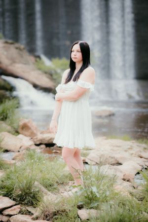 Person in white dress poses near waterfall in dreamy outdoor portrait setting.