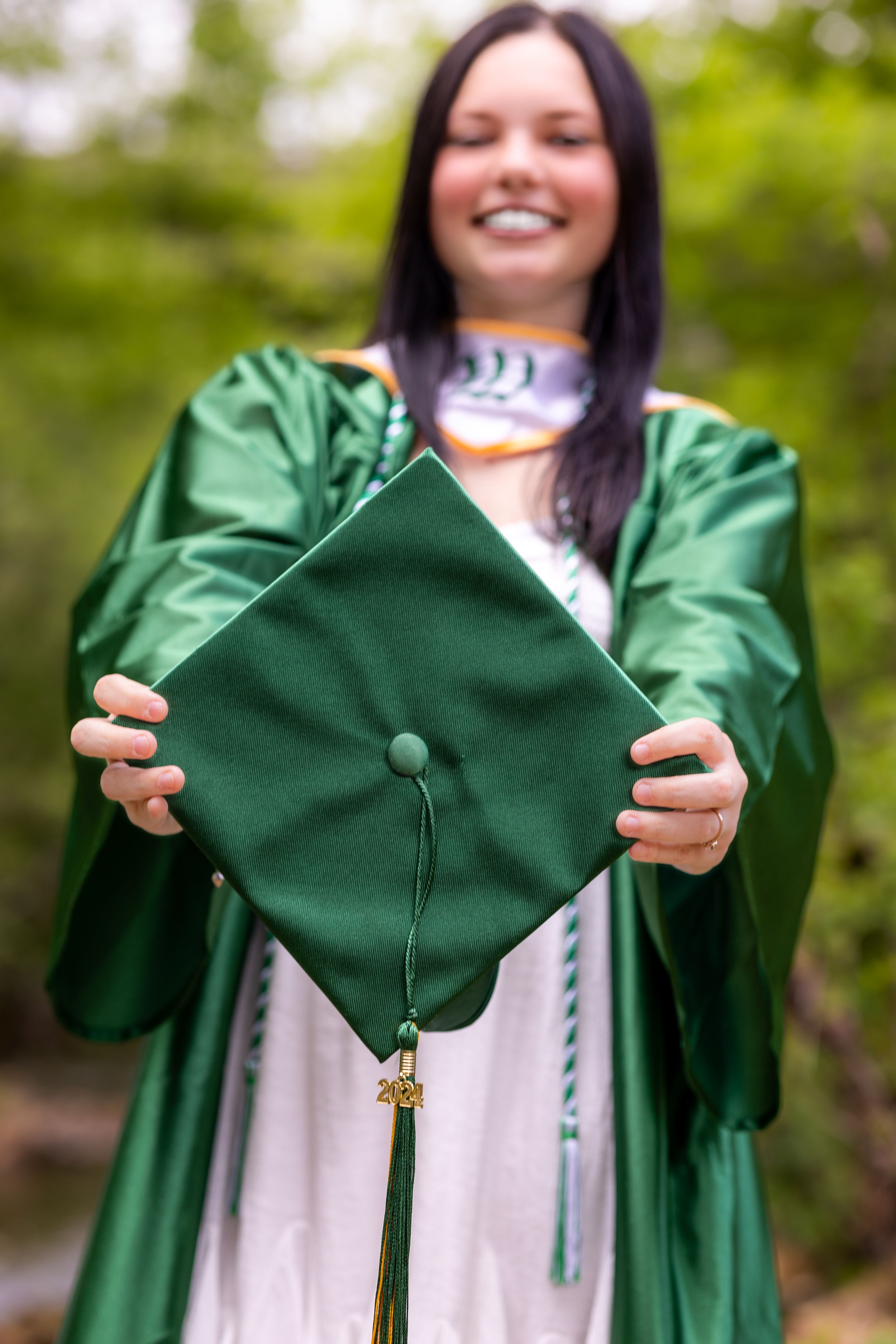 Graduate in green cap and gown holding up decorative graduation cap with tassel outdoors.