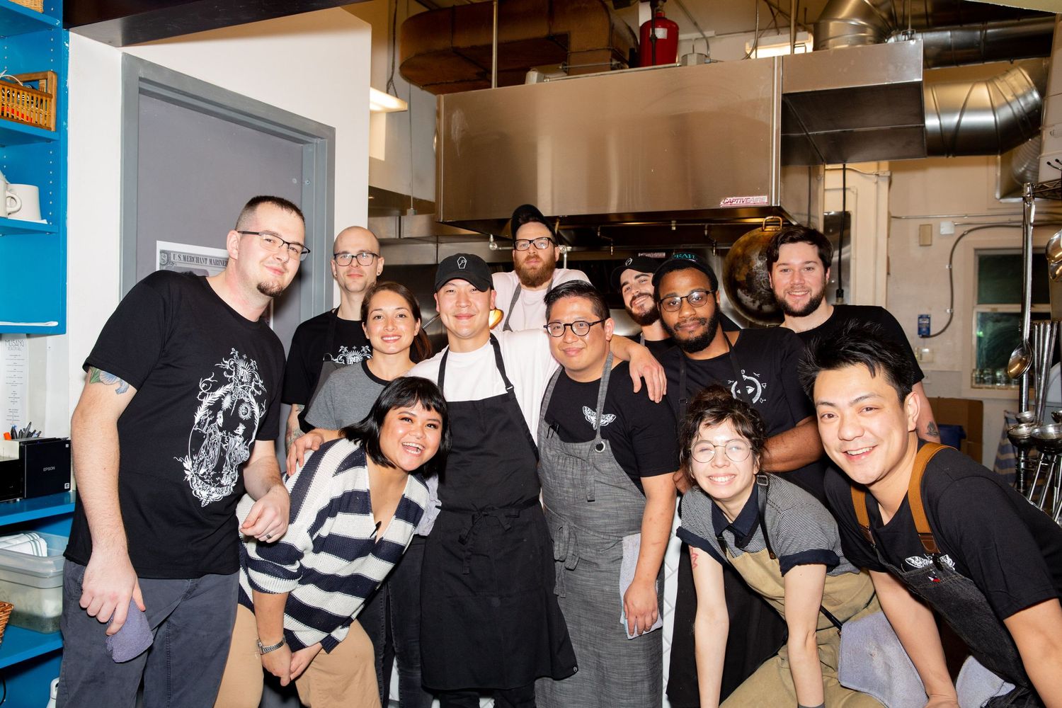 Restaurant kitchen staff members pose together for a group photo wearing aprons and casual attire.