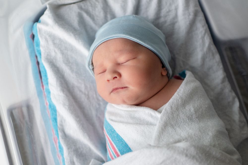 A sleeping newborn baby swaddled in white blankets wearing a light blue hat.