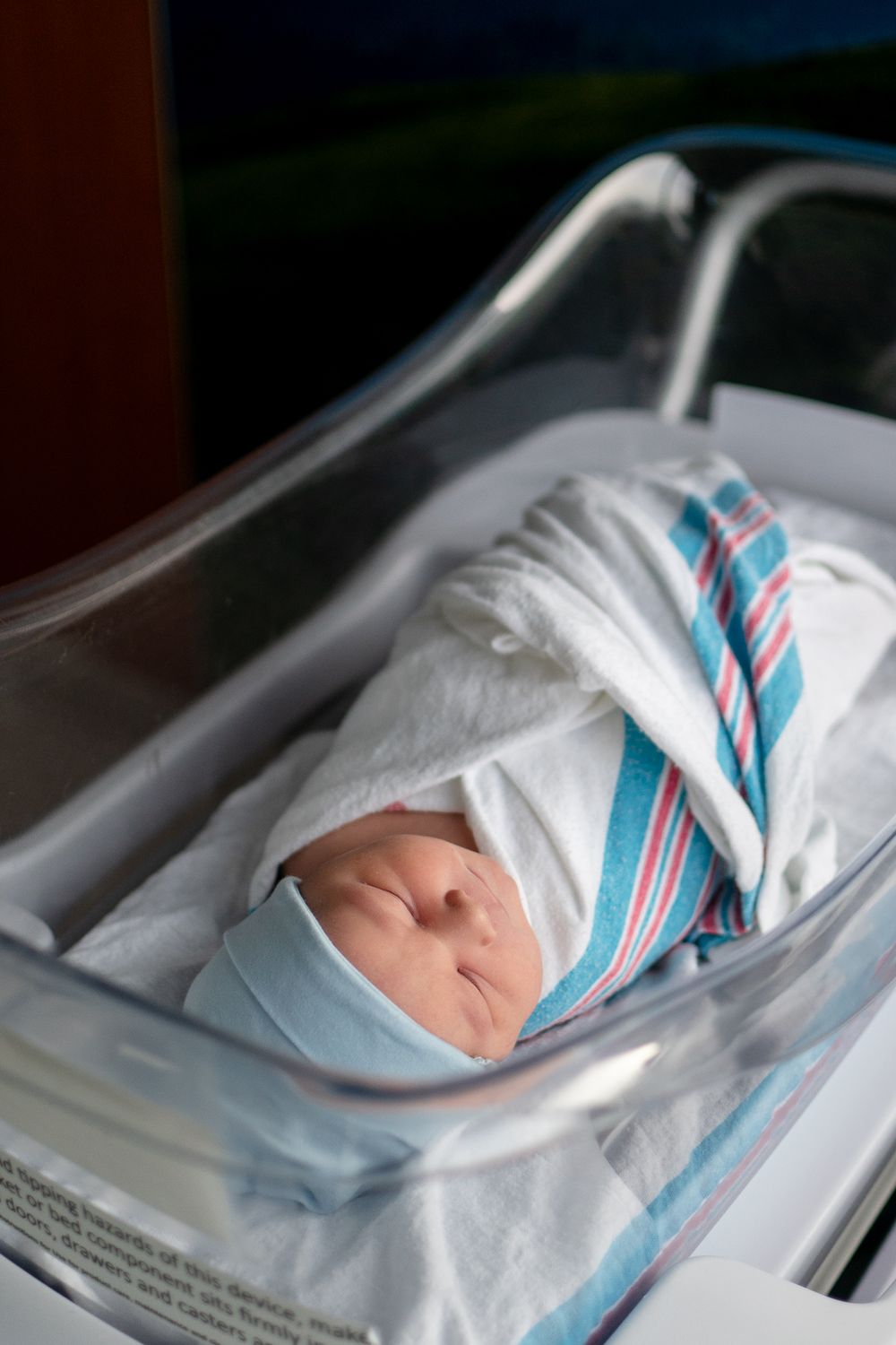 A newborn baby swaddled in white and blue striped hospital blanket rests in a bassinet.