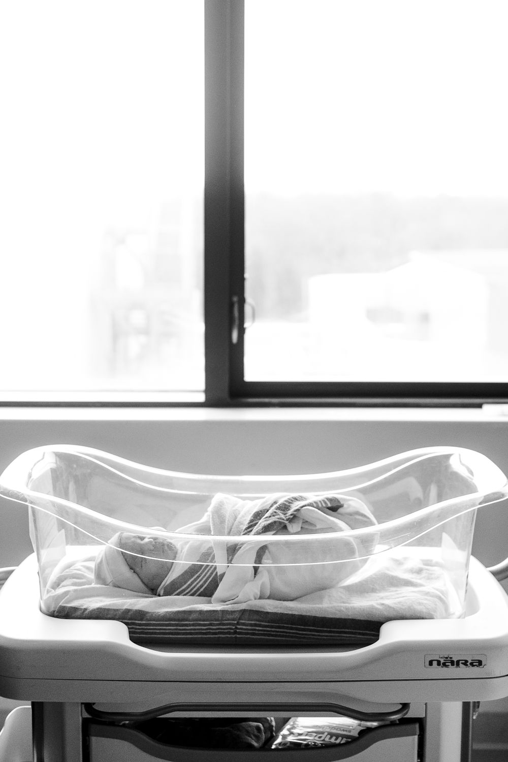 A newborn sleeps peacefully in a clear hospital bassinet in black and white photography.