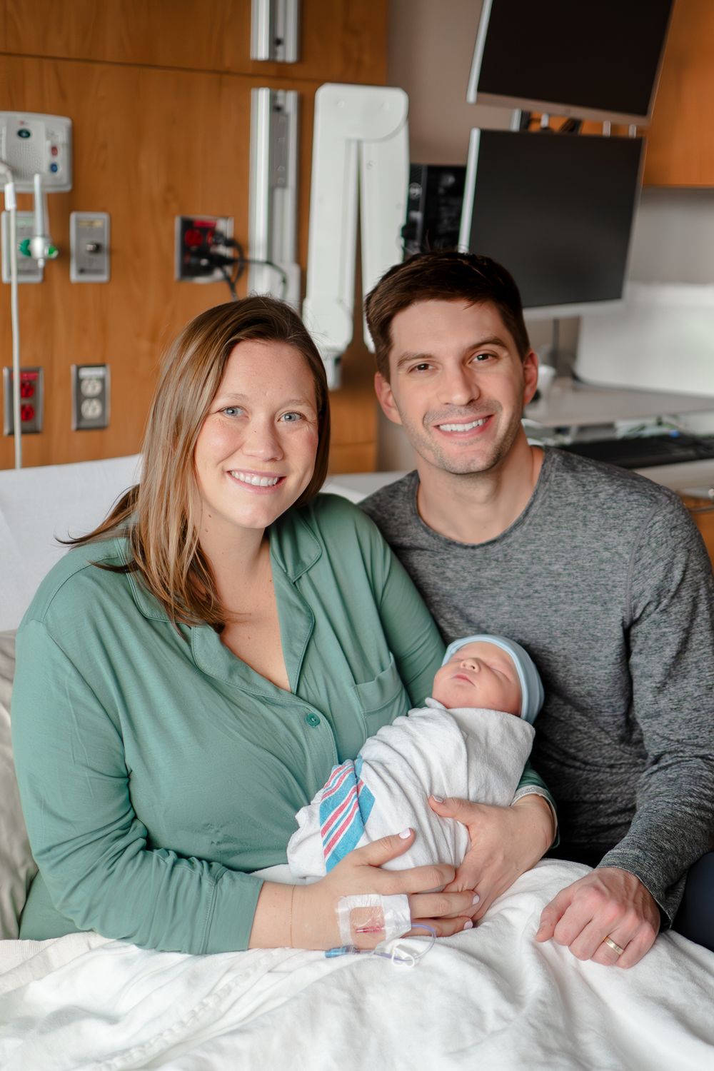 A couple smiles while holding their newborn baby in a hospital room with medical equipment visible in the background.