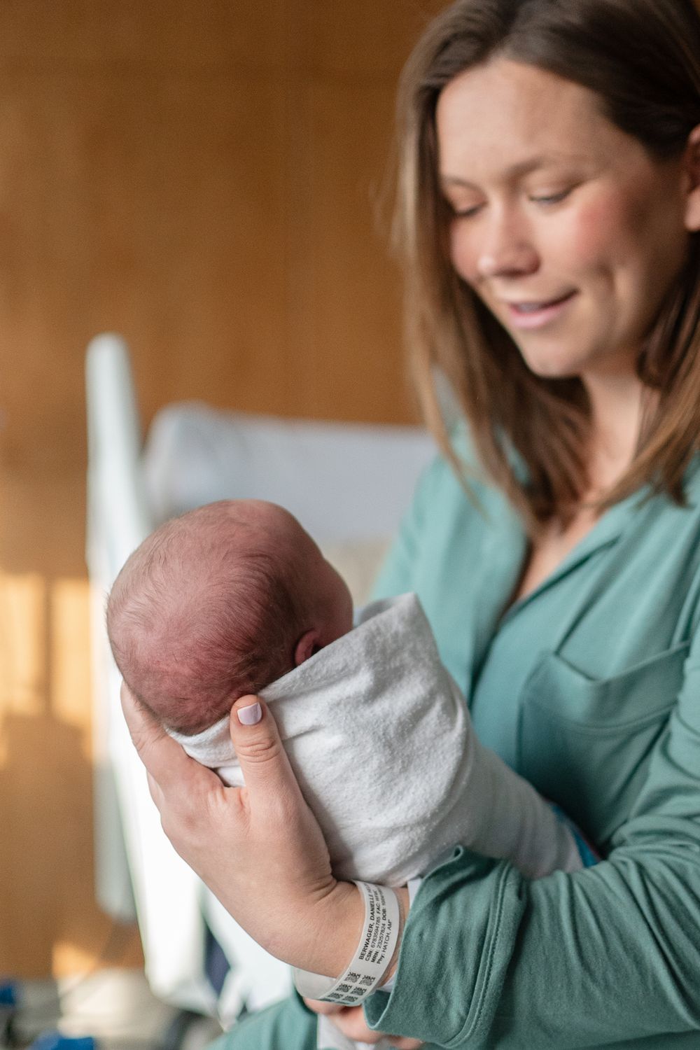 A newborn baby wrapped in a soft gray blanket is held by someone wearing a mint green sweater in warm natural light.