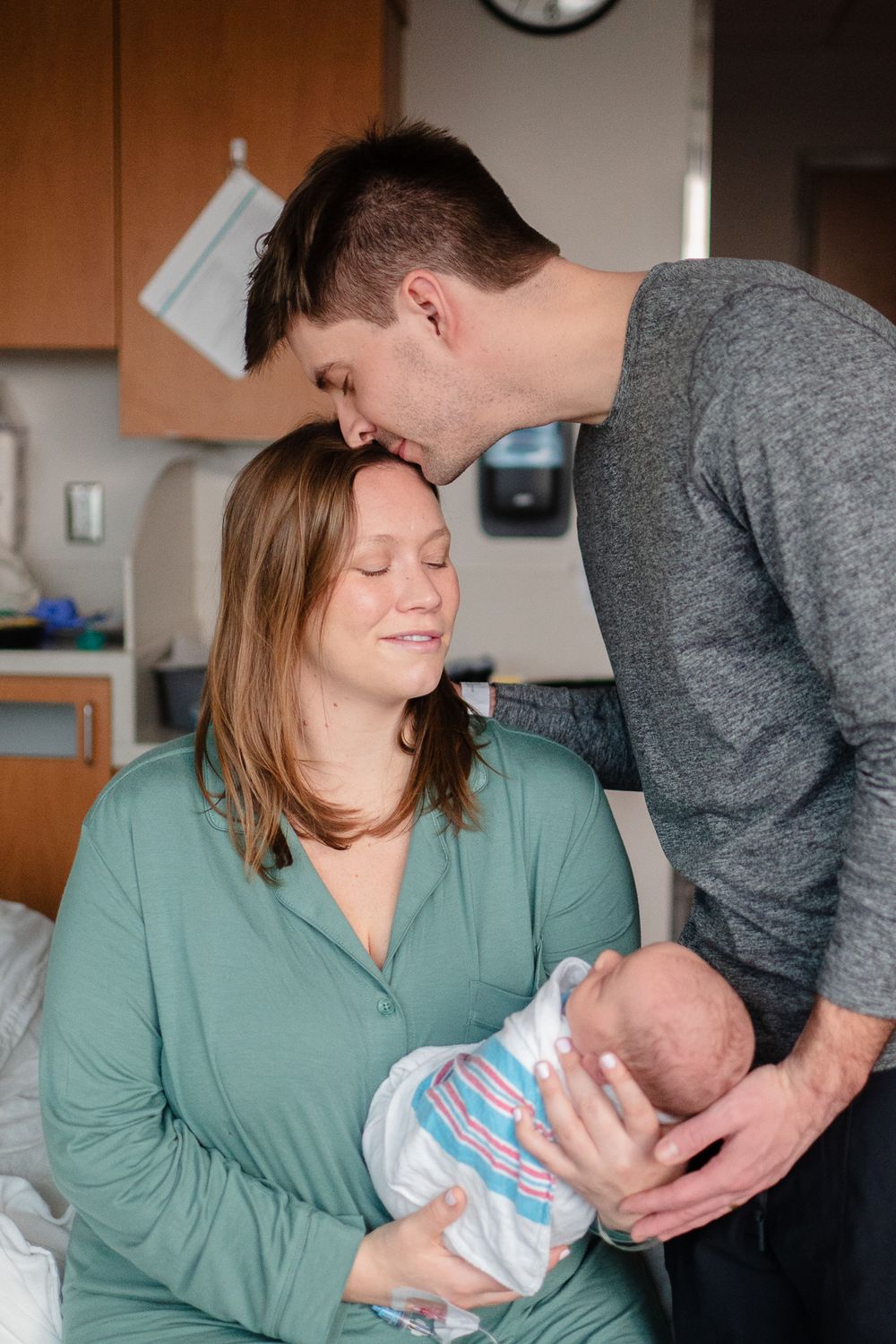 A tender moment as parents share their affection while holding their newborn baby wrapped in a striped blanket.