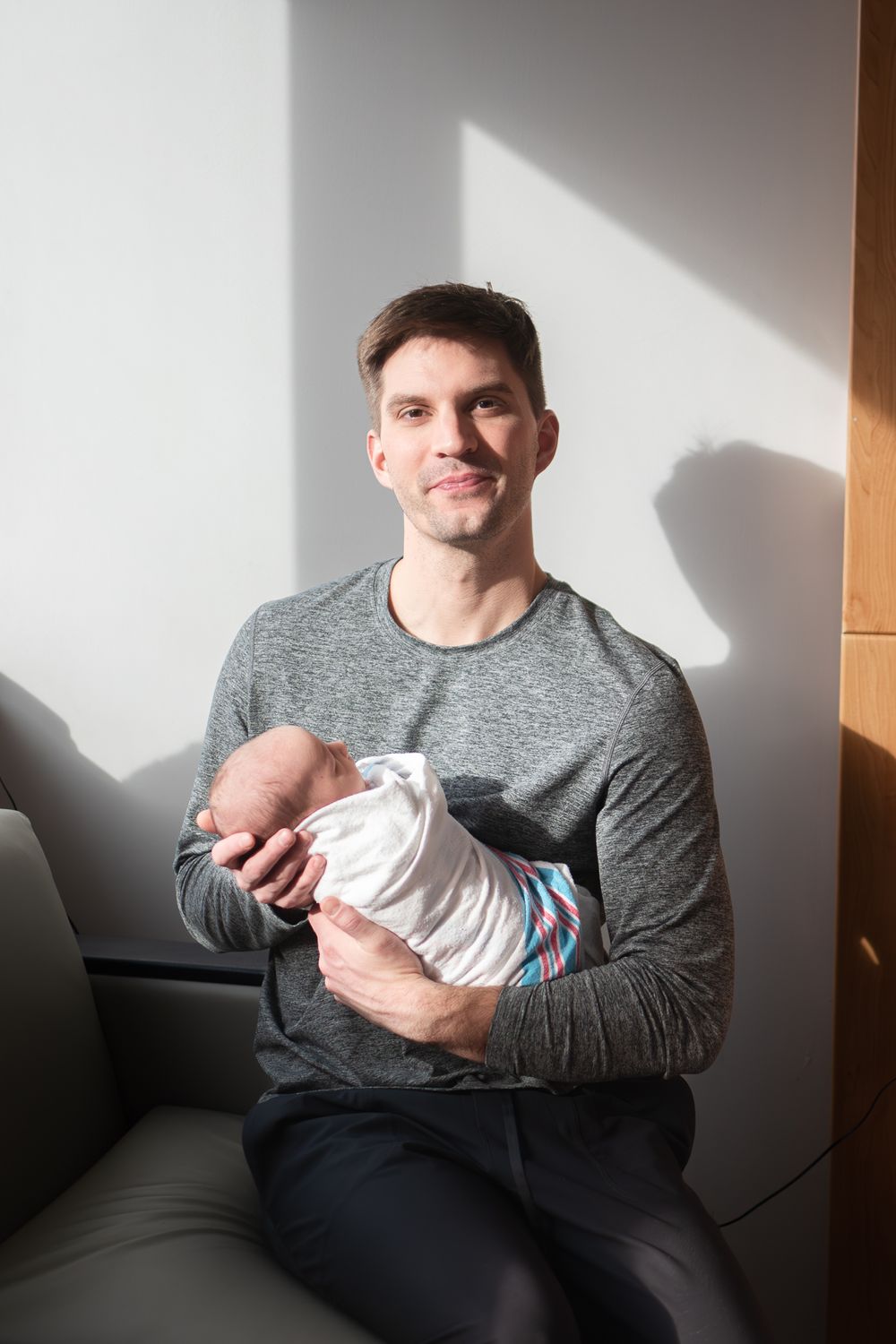 Person in grey sweater sits on couch holding small infant wrapped in white blanket as sunlight streams through window.