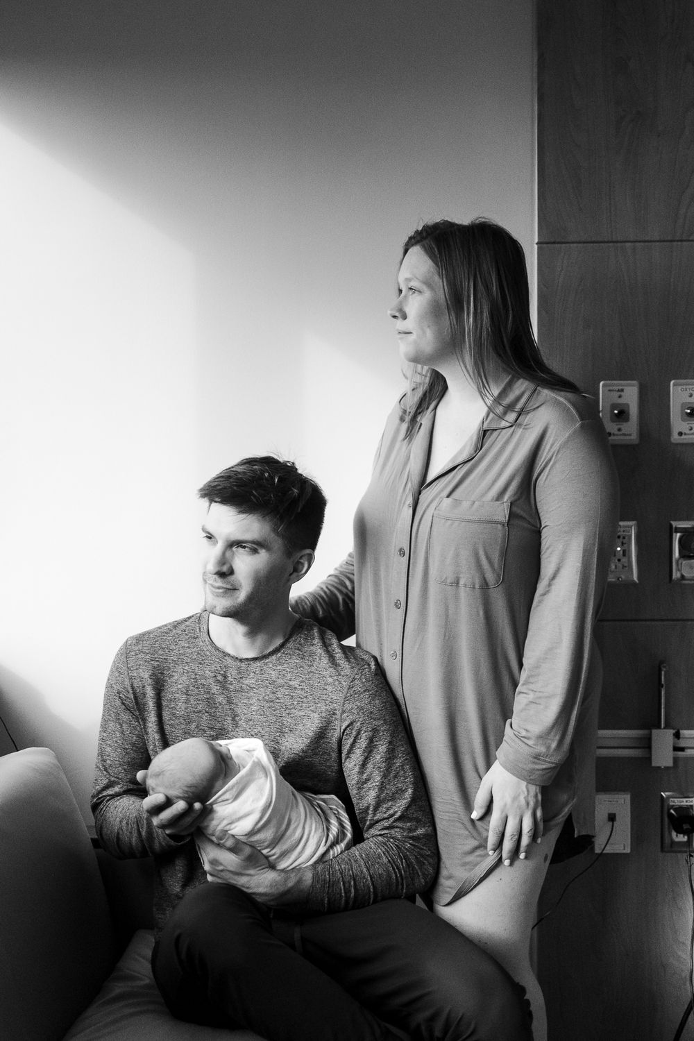 Black and white portrait of a couple in a relaxed indoor setting with dramatic side lighting through a window.