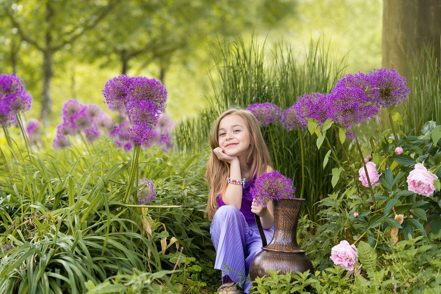 Kinderfotografie, Child in purple dress sits among blooming allium flowers and roses in garden.