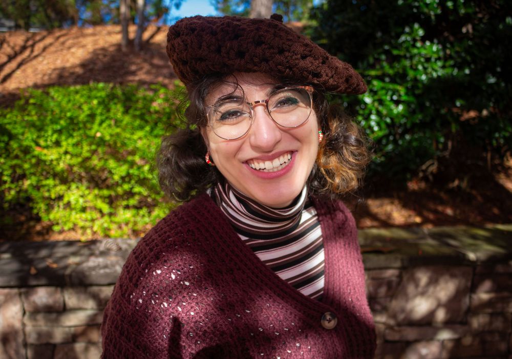 A person wearing a burgundy sweater and round glasses sits outdoors among green foliage on a sunny day.