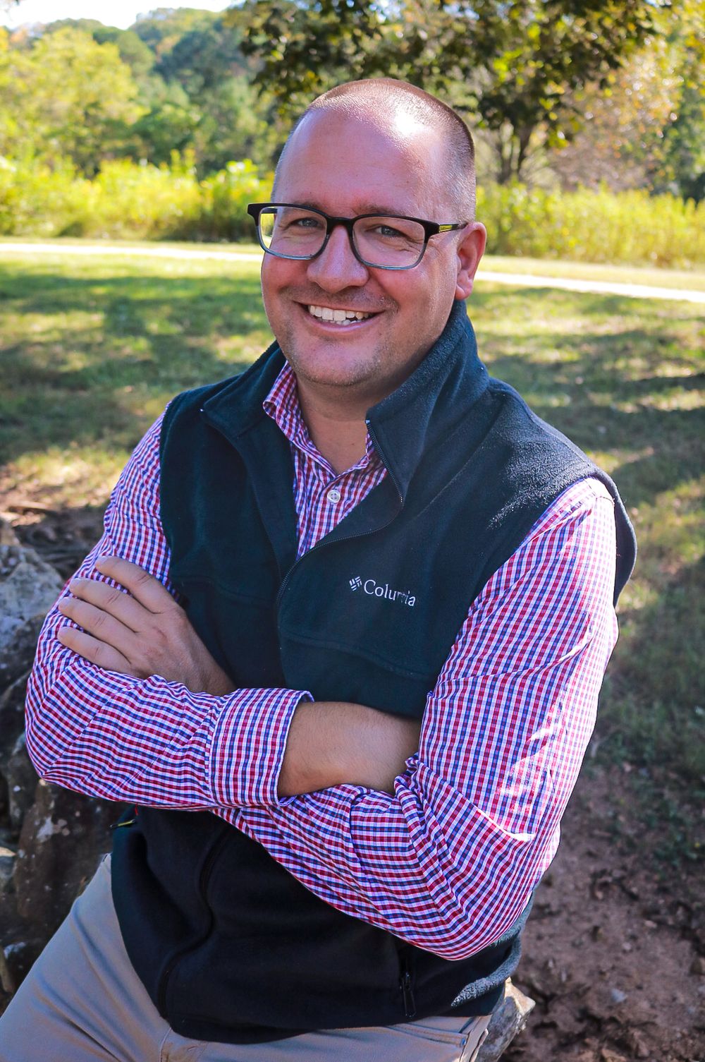 A person wearing a navy vest and red checkered shirt poses outdoors in a natural setting with trees and foliage.