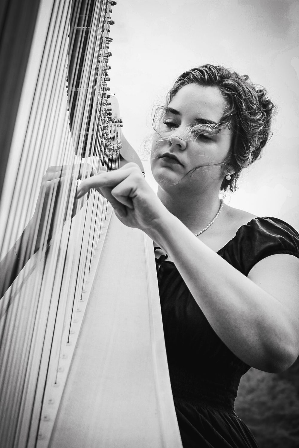 A classical musician plays a harp in black and white, wearing a dark dress and delicate necklace.