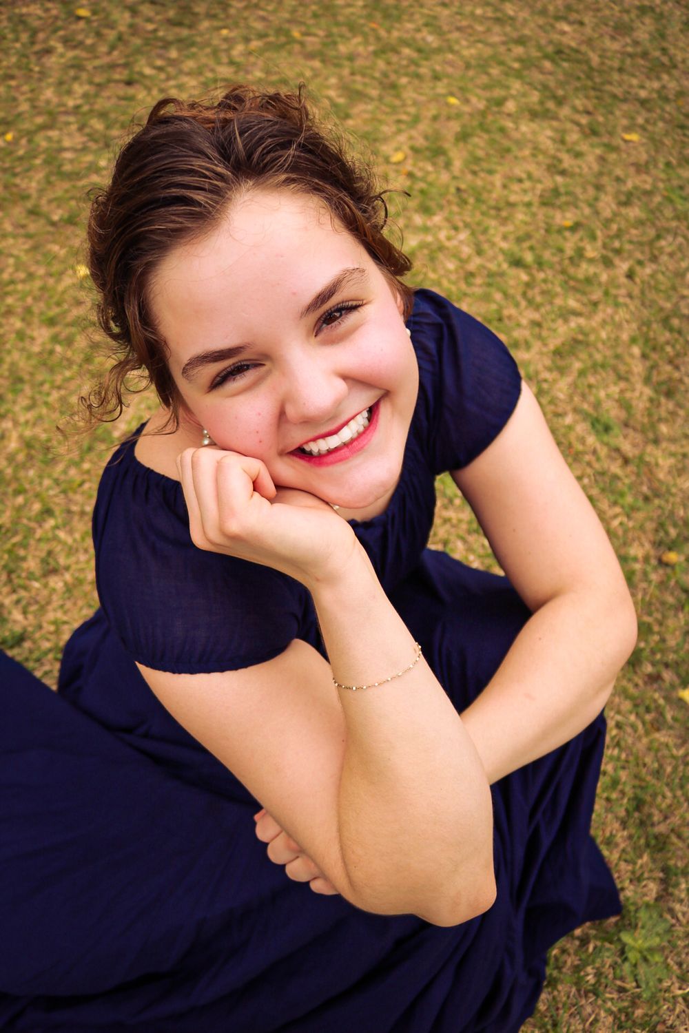 Aerial view of someone in a navy blue dress sitting on grass with a warm smile while resting their chin on their hand.