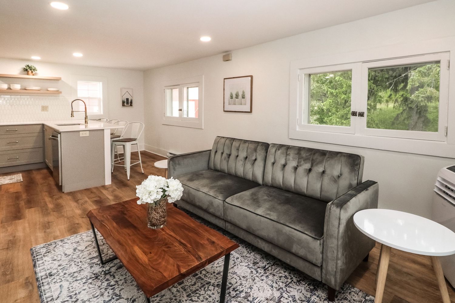 Modern living room with gray tufted sofa, wood coffee table, hardwood floors, and open kitchen view.