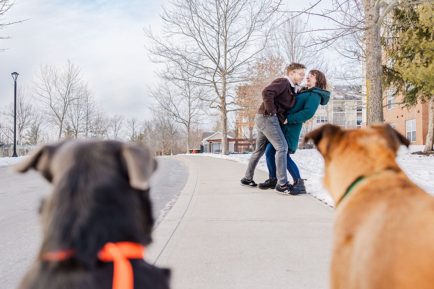 Two people embrace outdoors in winter while their dogs watch in the foreground.