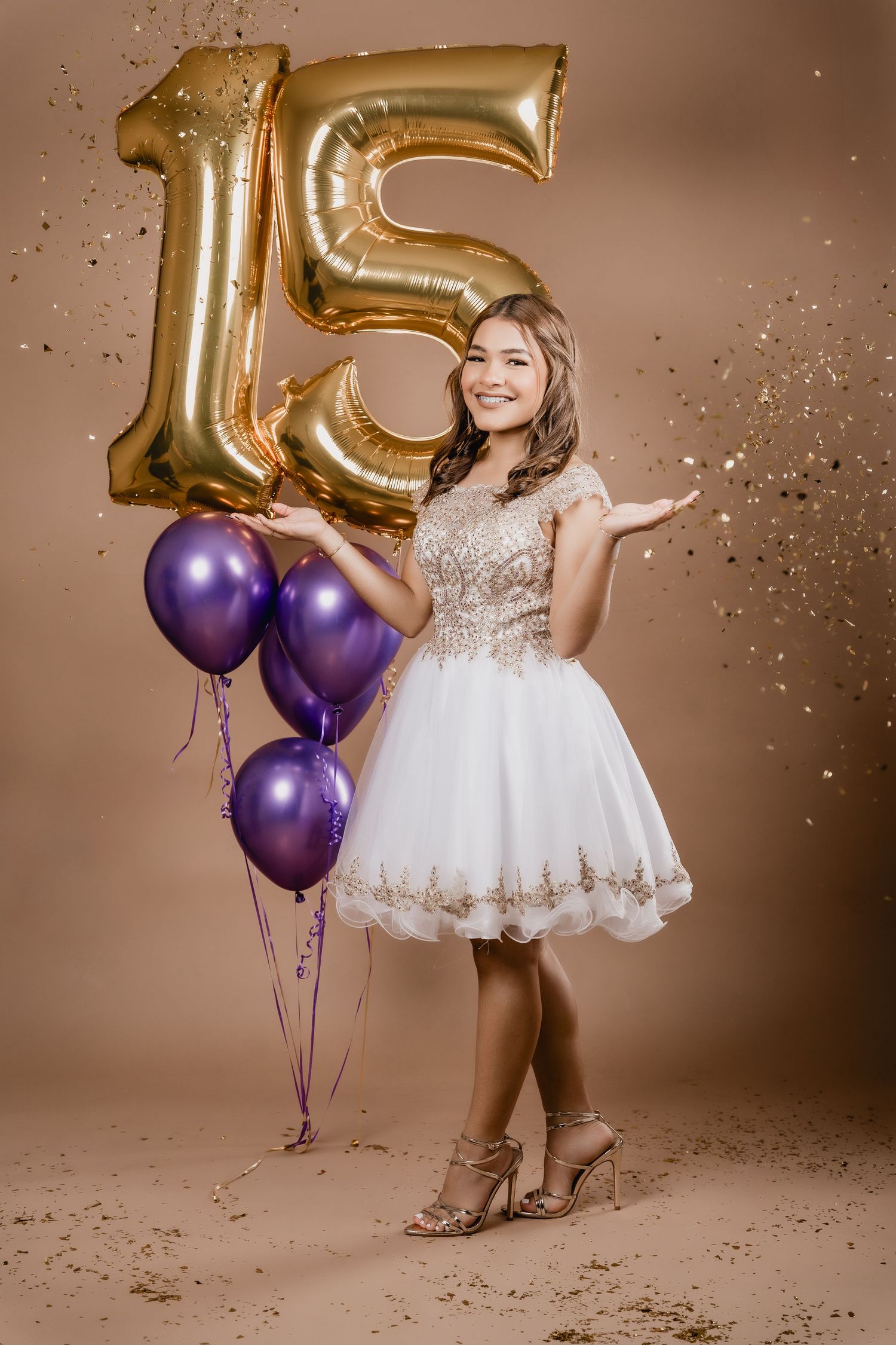 A celebratory scene with gold number 15 balloons, purple balloons, and a white and gold party dress against a brown background.