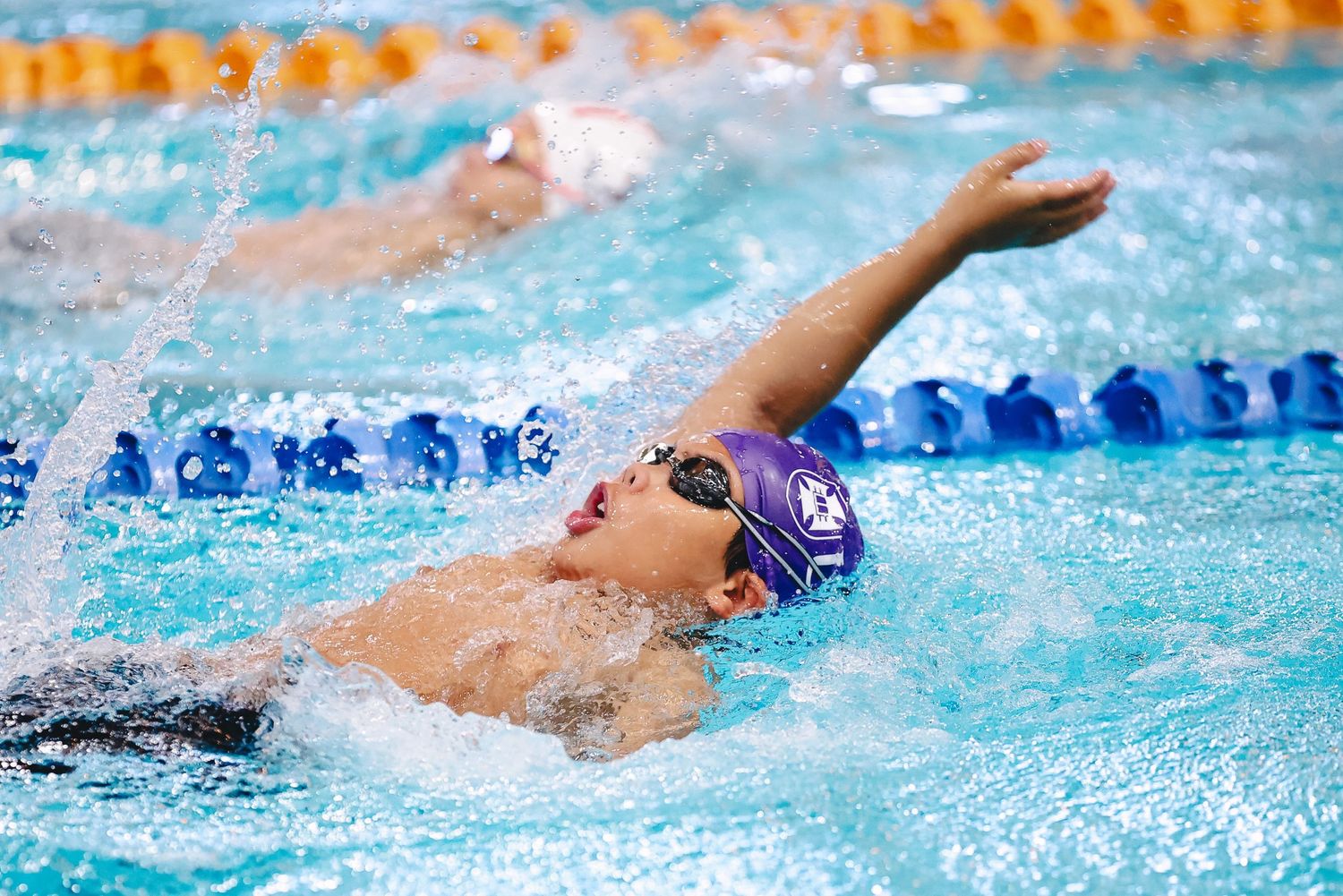 Competitive swimmer in purple cap performs backstroke during race in swimming pool with lane dividers.