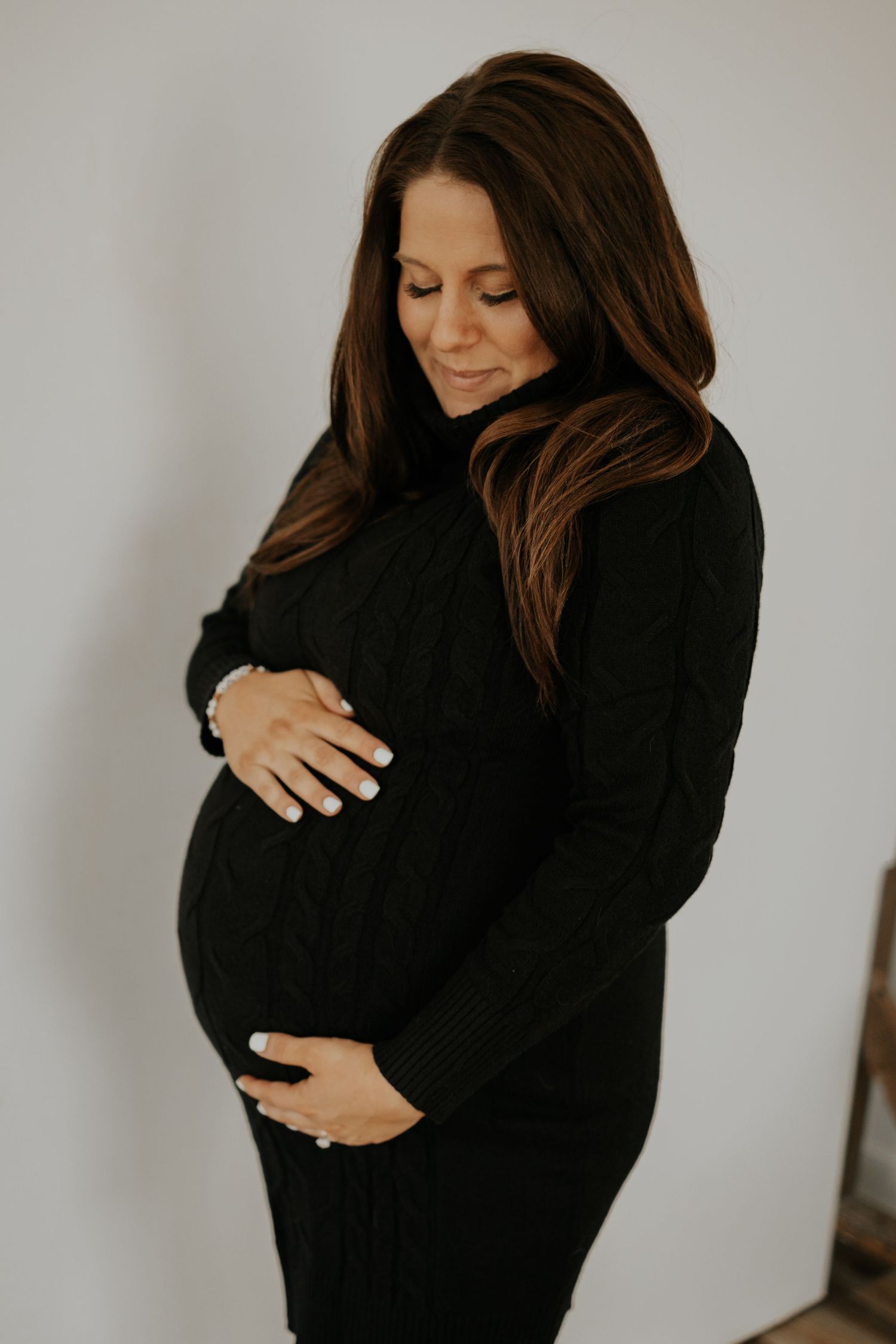 A pregnant woman in a black sweater dress lovingly holds her baby bump against a neutral background.