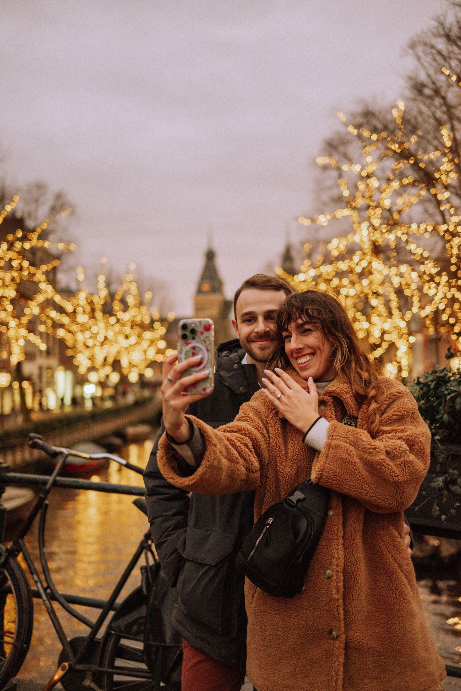 A couple in Amsterdam during winter, standing close and smiling against twinkling lights after an engagement proposal.