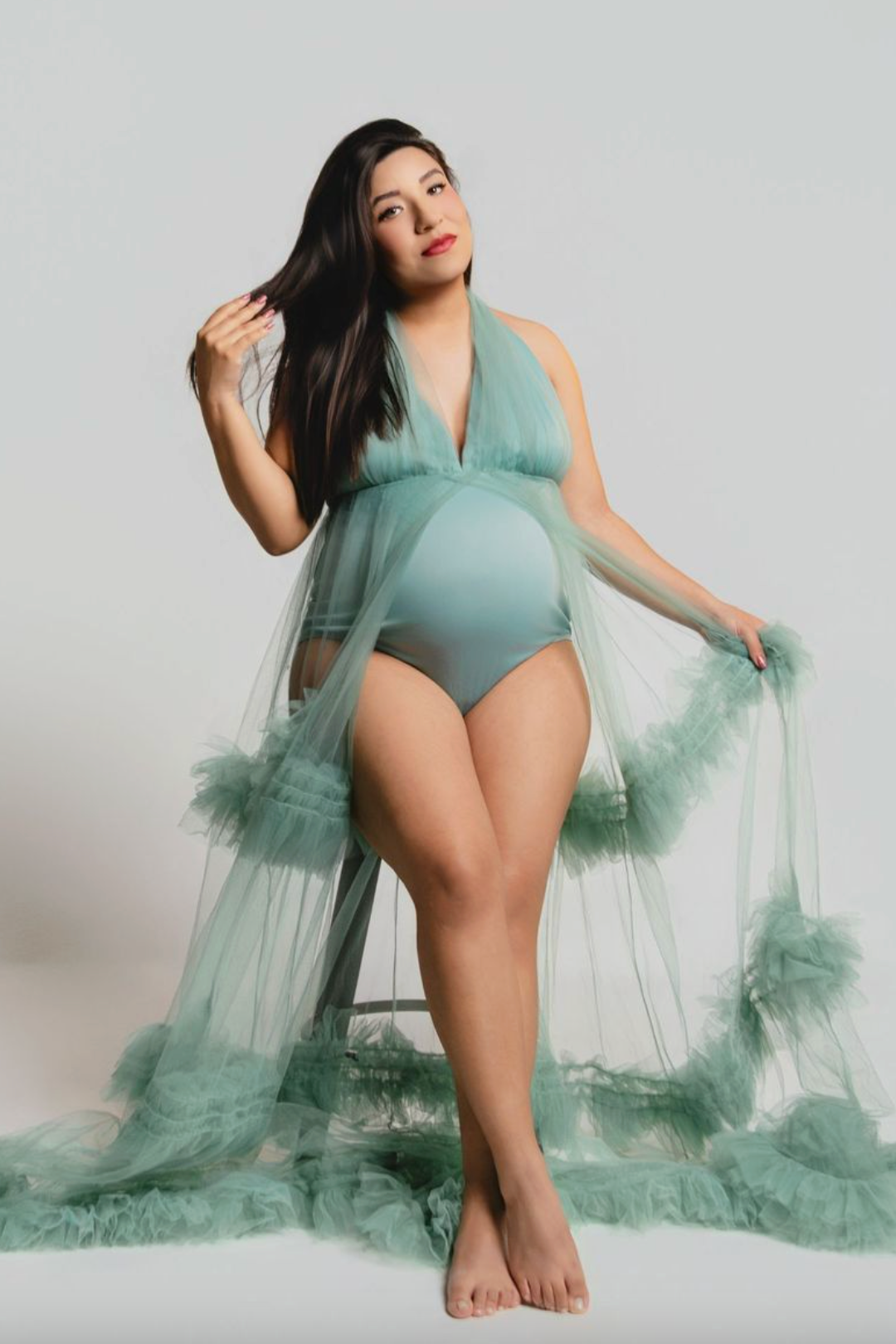 Pregnant woman in teal bodysuit and sheer ruffled robe poses gracefully in a professional maternity photo shoot.