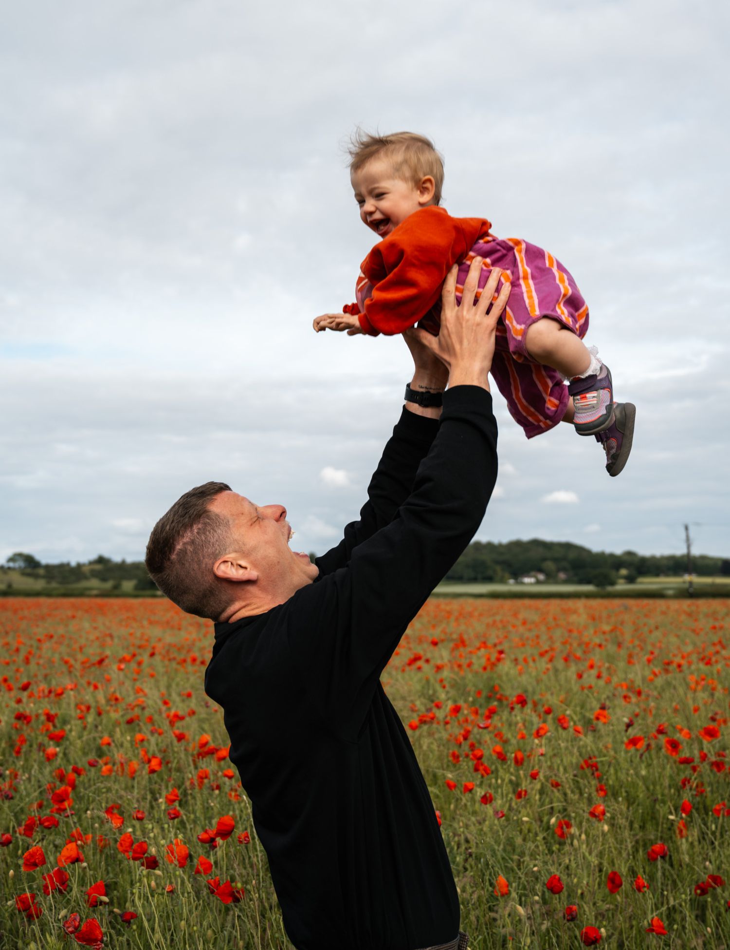 Father joyfully lifting laughing toddler above a vibrant red poppy field.