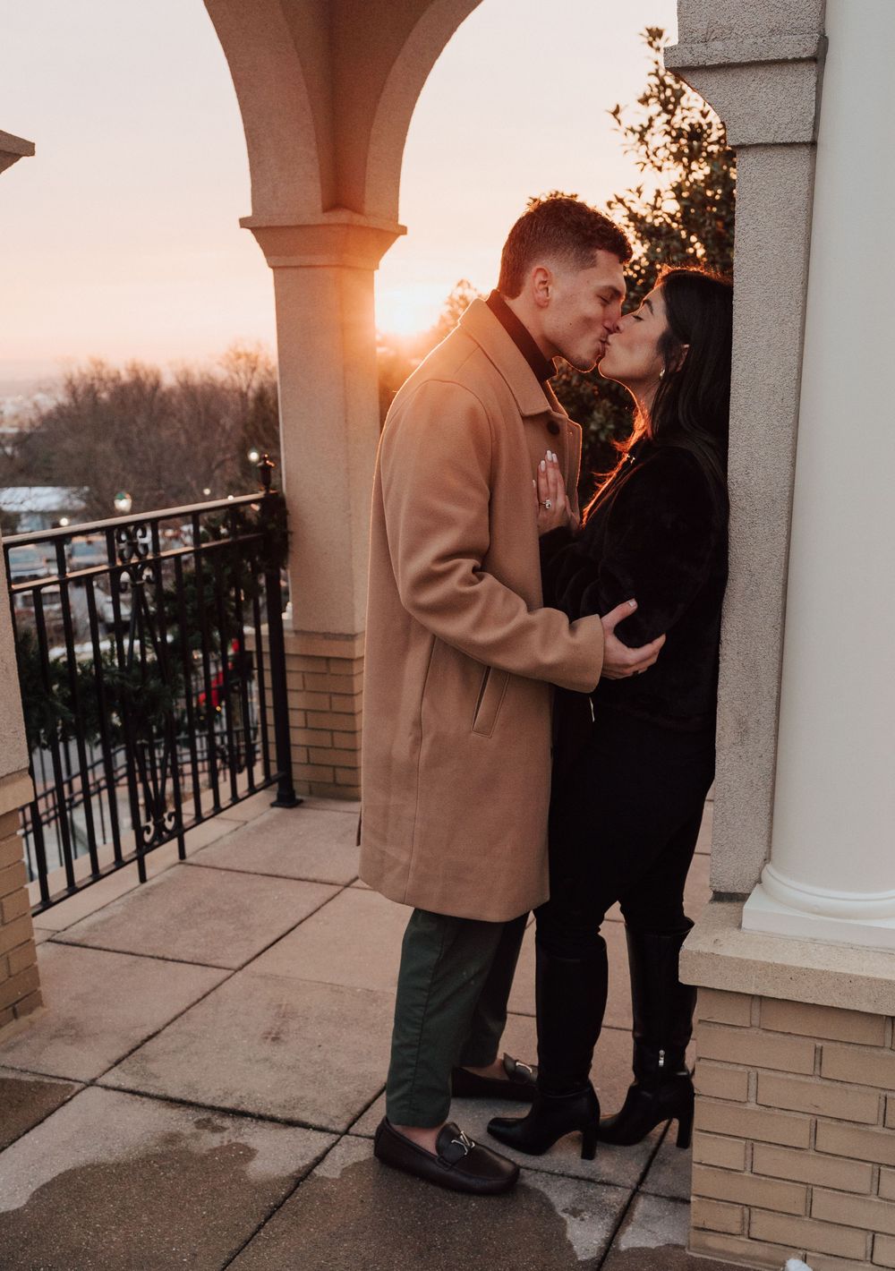Couple sharing a romantic kiss at sunset on an elegant balcony with arched columns and city views.