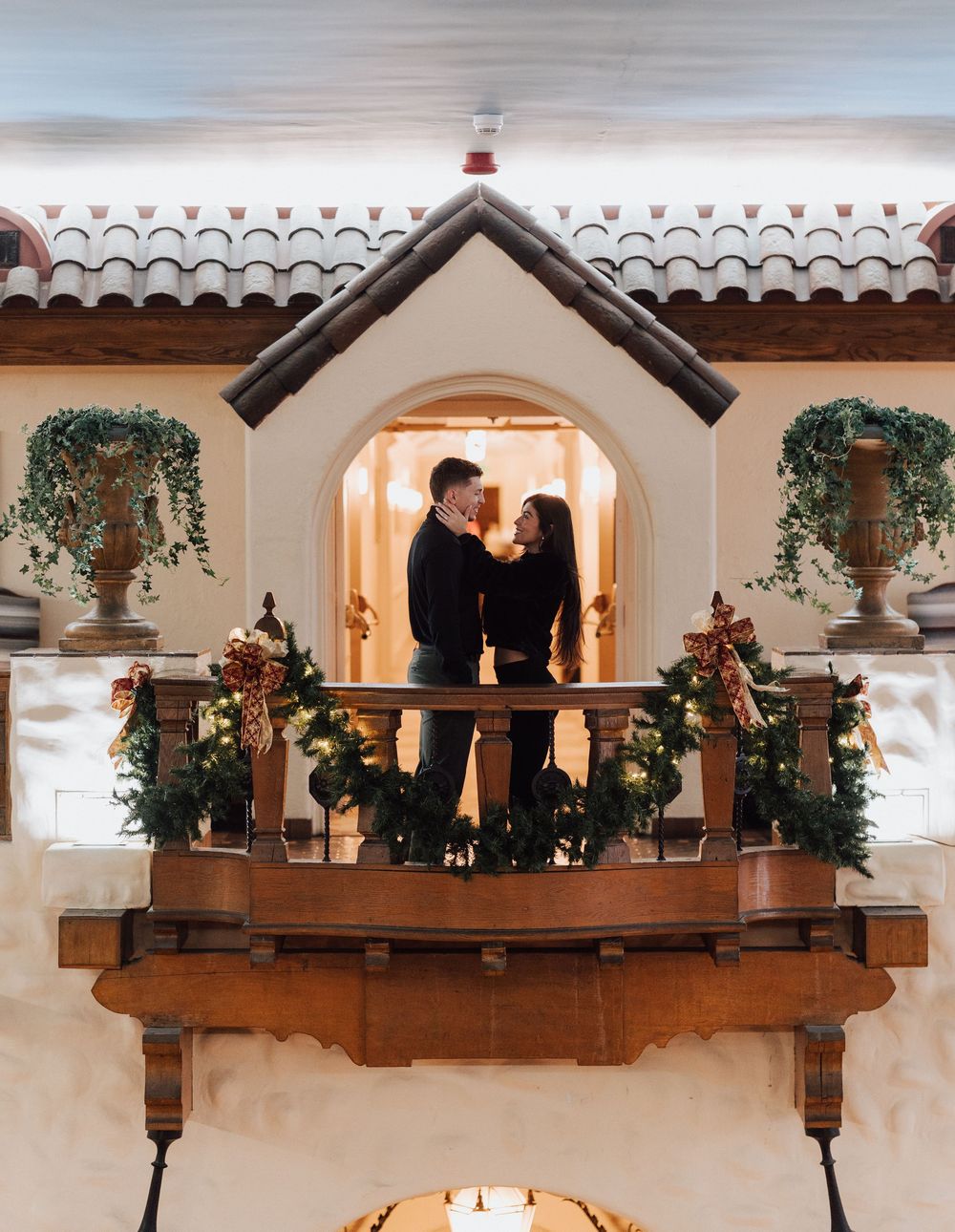 Couple embracing on a decorated balcony with holiday garland and lights in an elegant Spanish-style venue.