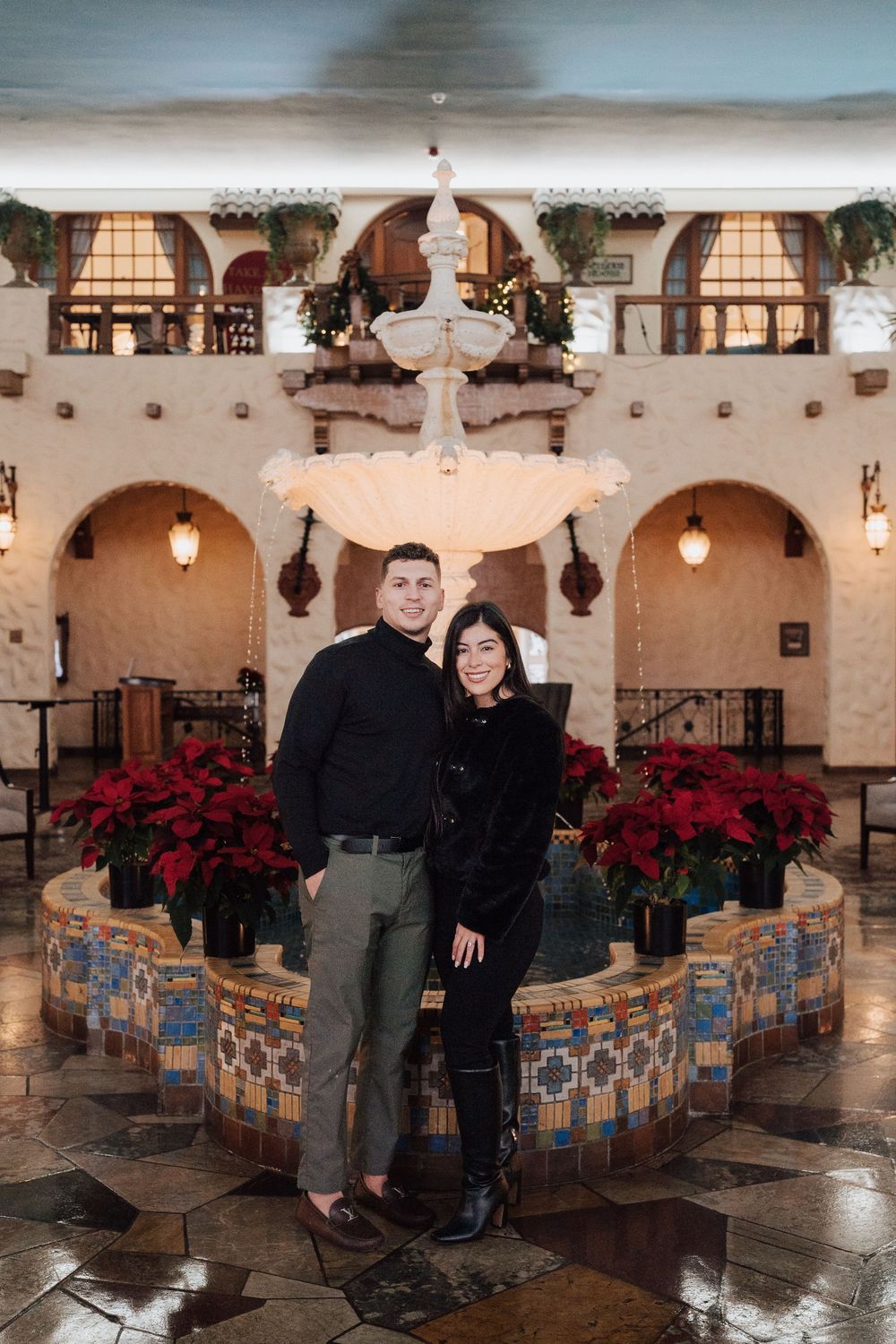 Couple smiling together in front of a decorative fountain surrounded by red poinsettias in a festive holiday lobby.