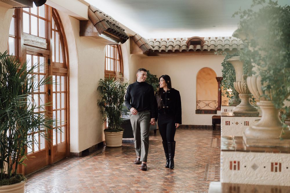 Couple walking hand in hand through elegant Spanish-style hotel corridor with arched windows and lush plants.