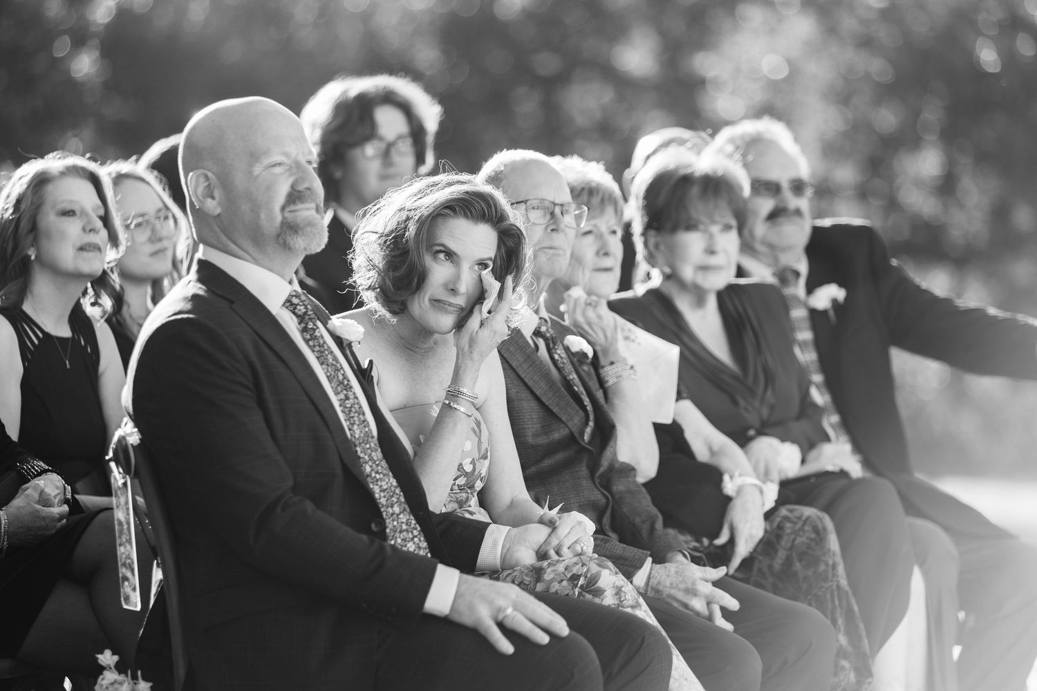 Black and white photo of wedding guests seated outdoors, smiling and watching a ceremony.