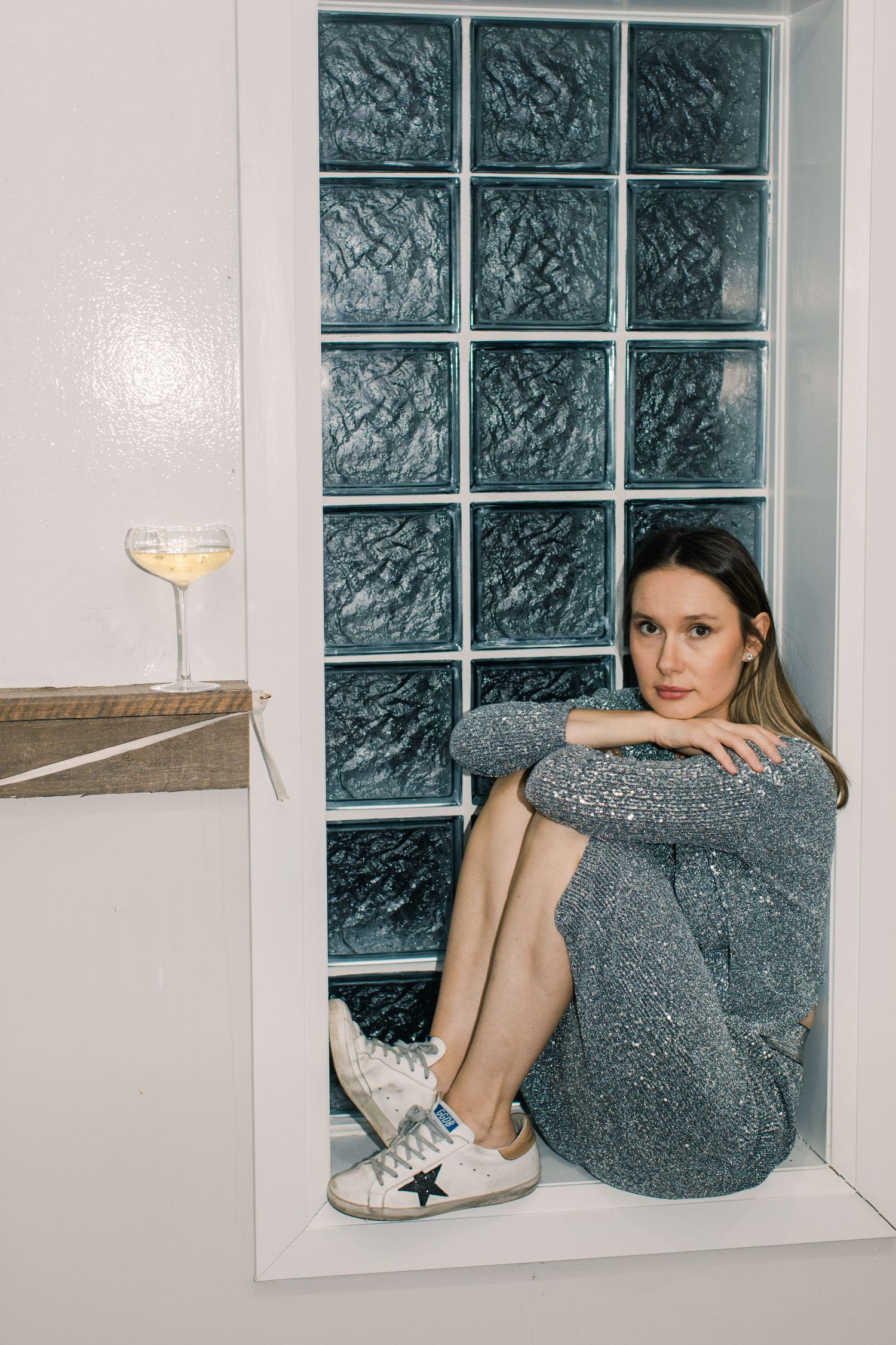 Woman in silver sequin dress and white star sneakers sitting in a glass block window niche with a wine glass nearby.