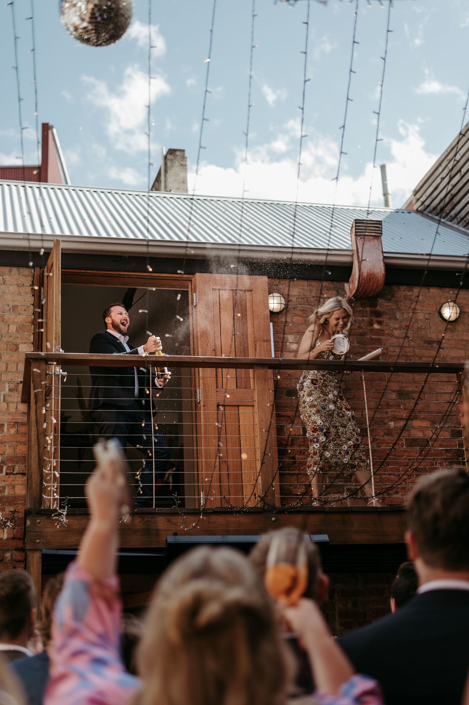 Couple celebrating on a balcony popping champagne at a wedding reception with guests cheering below.