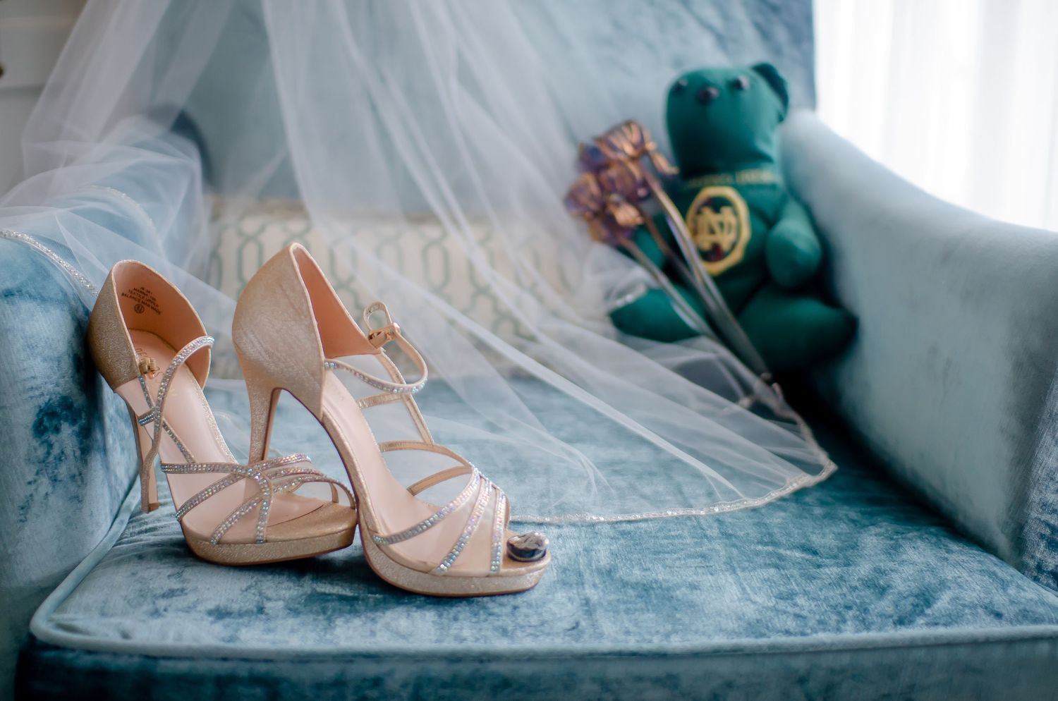 Elegant bridal heels with ankle straps rest on a blue velvet chair beside a wedding veil and green teddy bear.