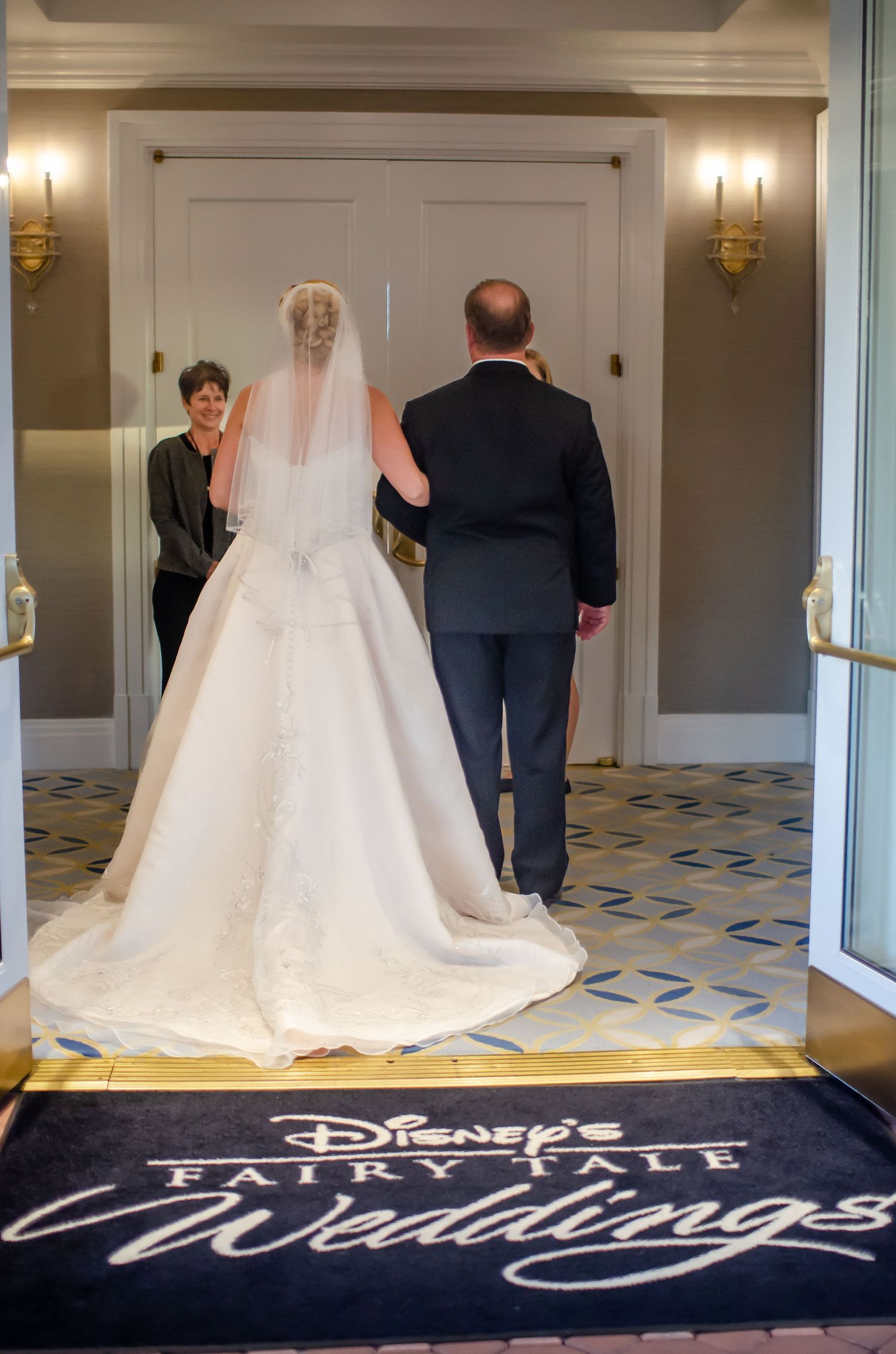 Bride in white gown and veil walks with escort on Disney's Fairytale Weddings carpet toward ceremony doors.