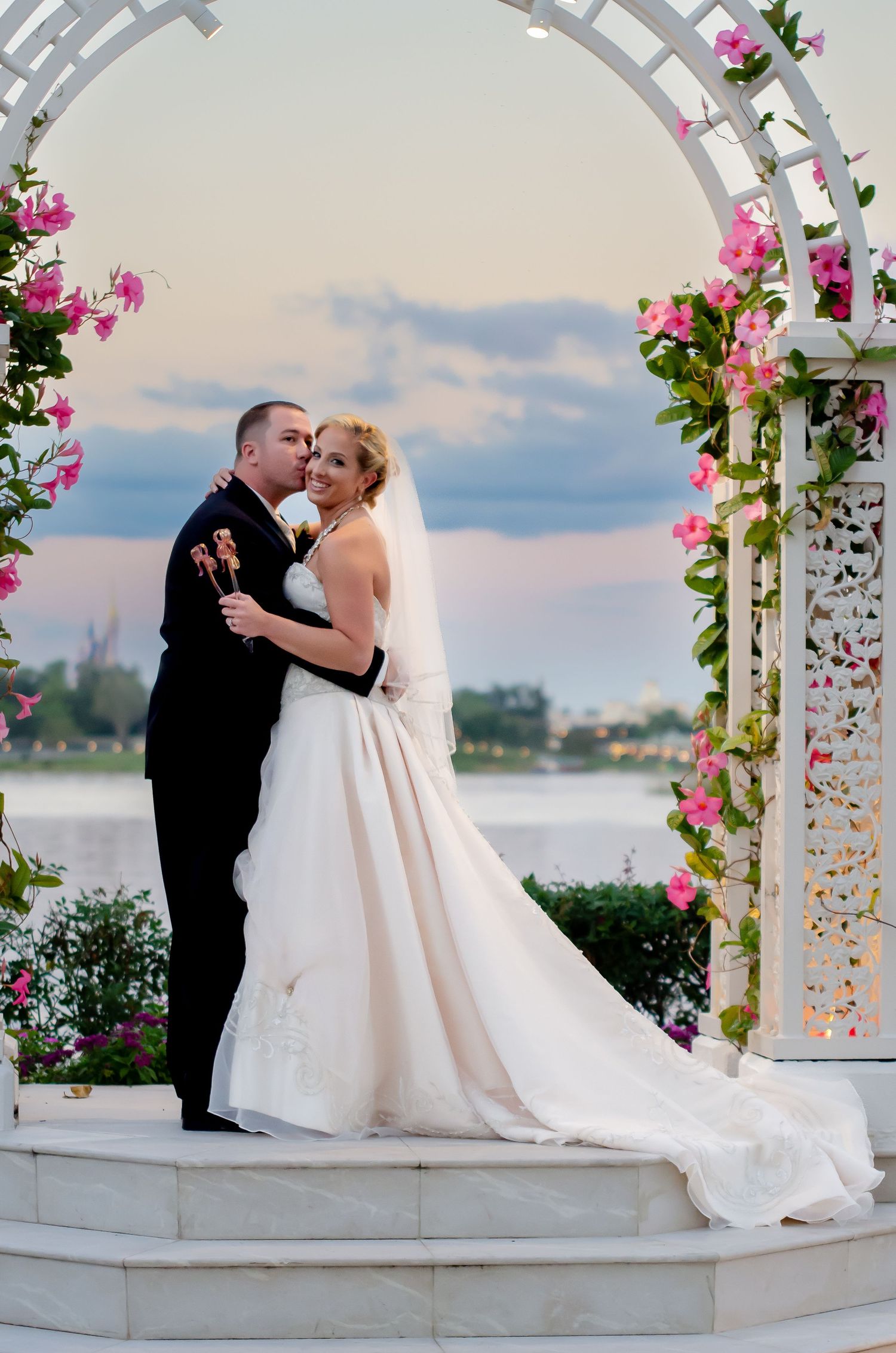 Bride and groom embrace under a white floral arch adorned with pink flowers, with a waterfront sunset backdrop.