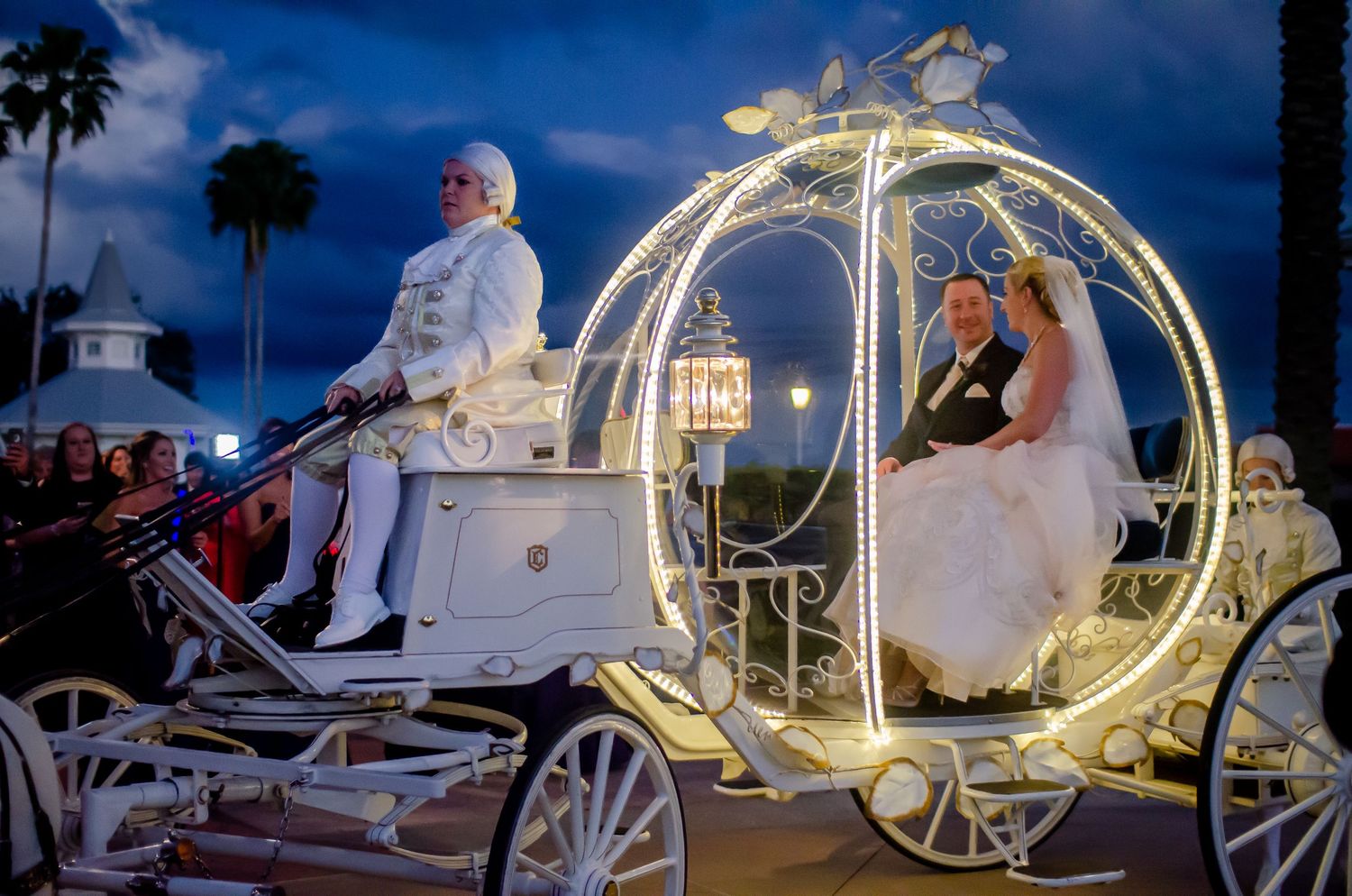 Bride and groom ride in an illuminated white Cinderella carriage at a fairy-tale wedding celebration at dusk.