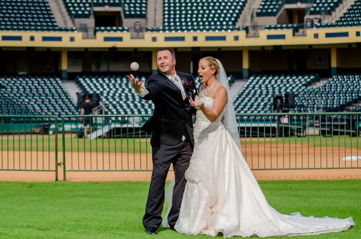Bride and groom posing on a baseball field in wedding attire, groom tossing a baseball in an empty stadium.