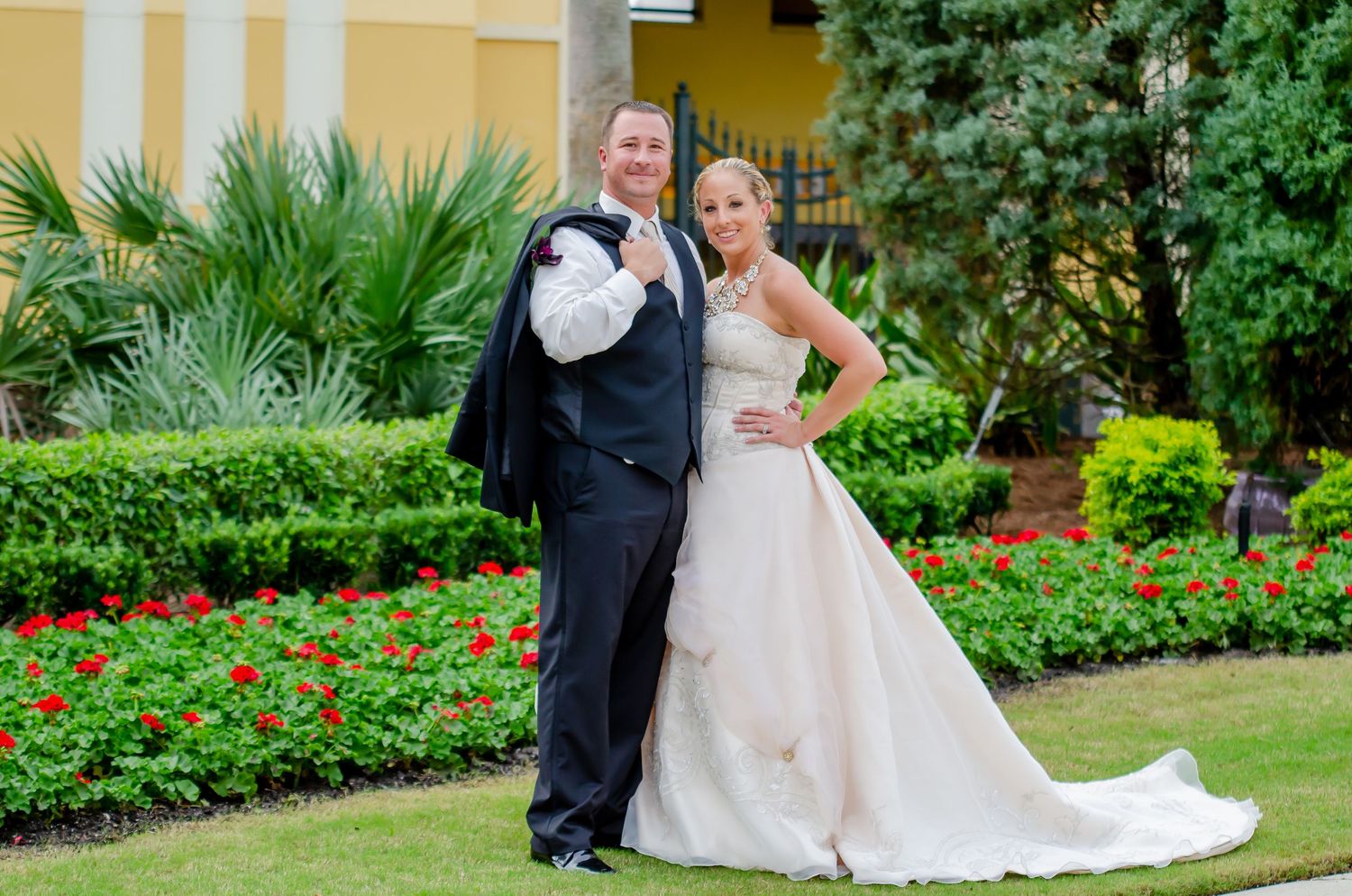 Bride in white gown and groom in black tuxedo posing together in a lush garden with red flowers.