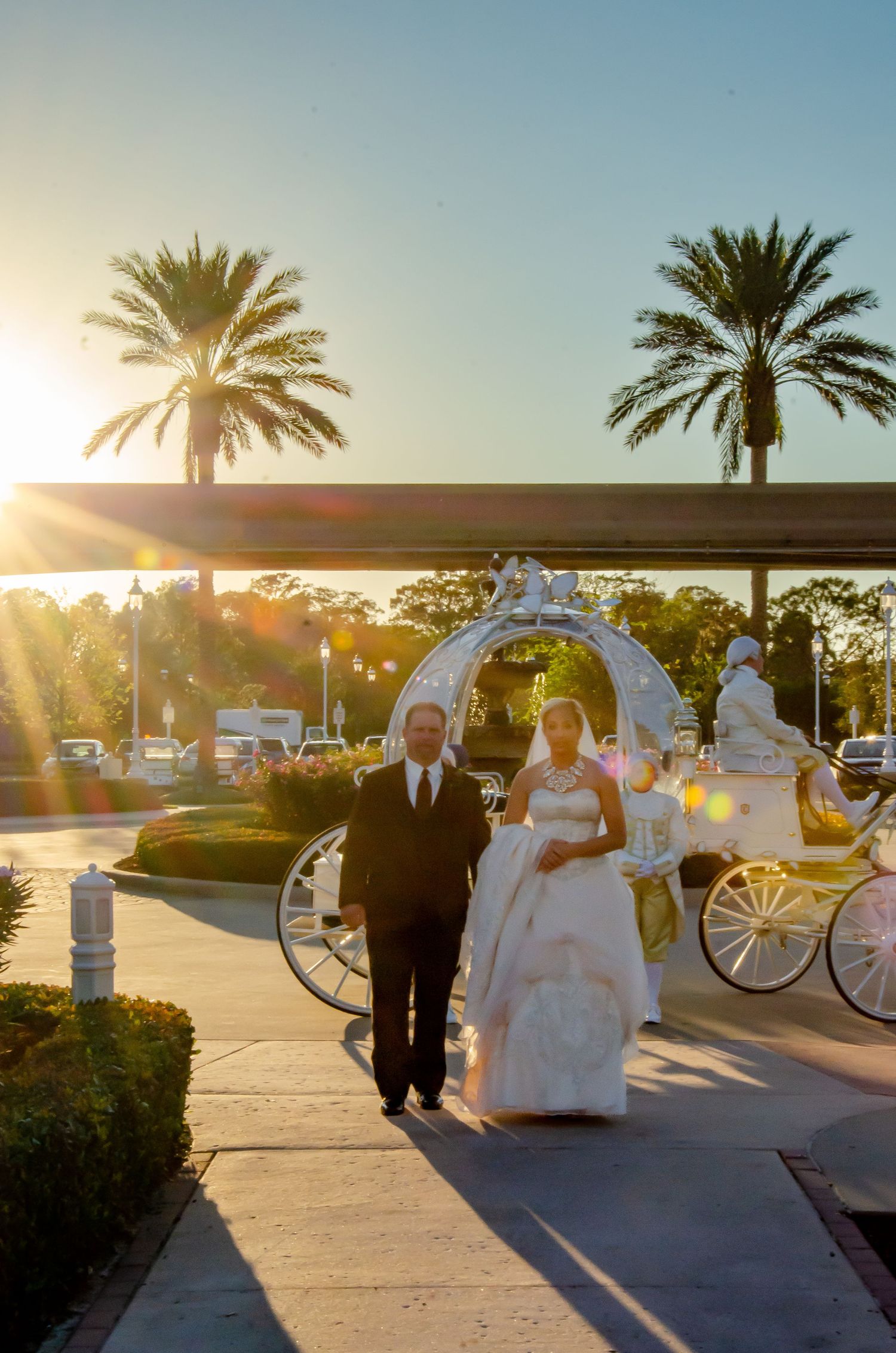 Bride and groom walk from a white Cinderella carriage at sunset with palm trees at a Florida wedding venue.