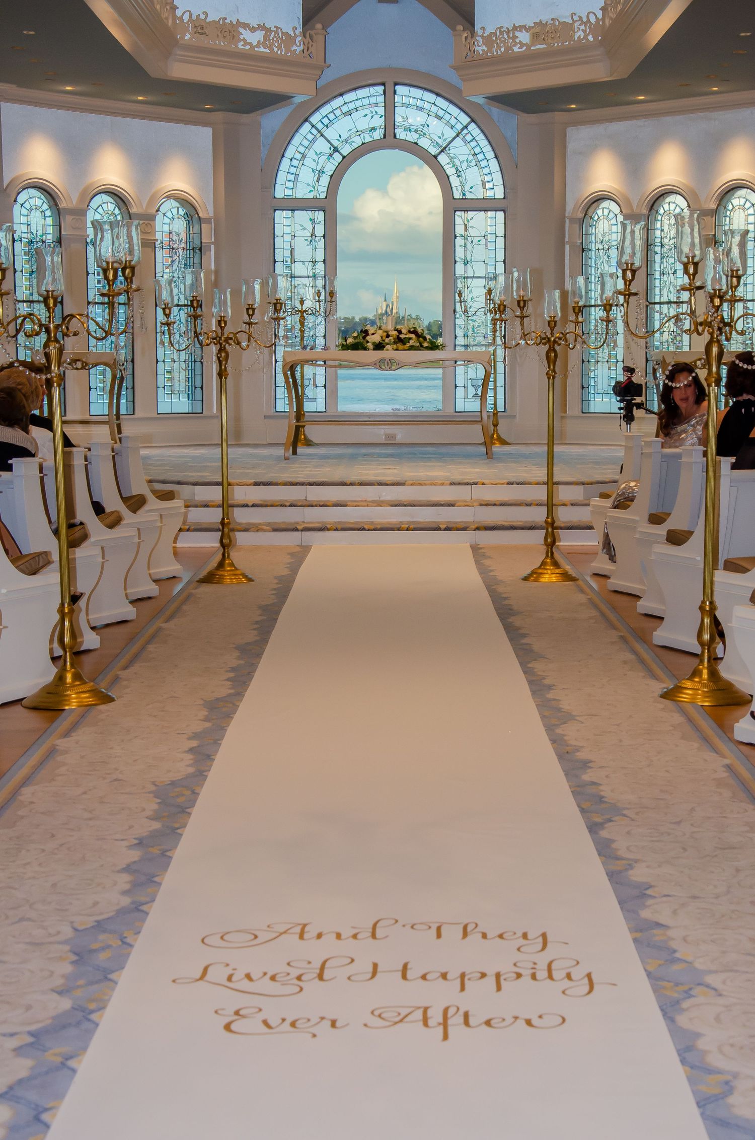 Elegant wedding chapel aisle with white runner reading And They Lived Happily Ever After, gold candelabras, and ocean view.