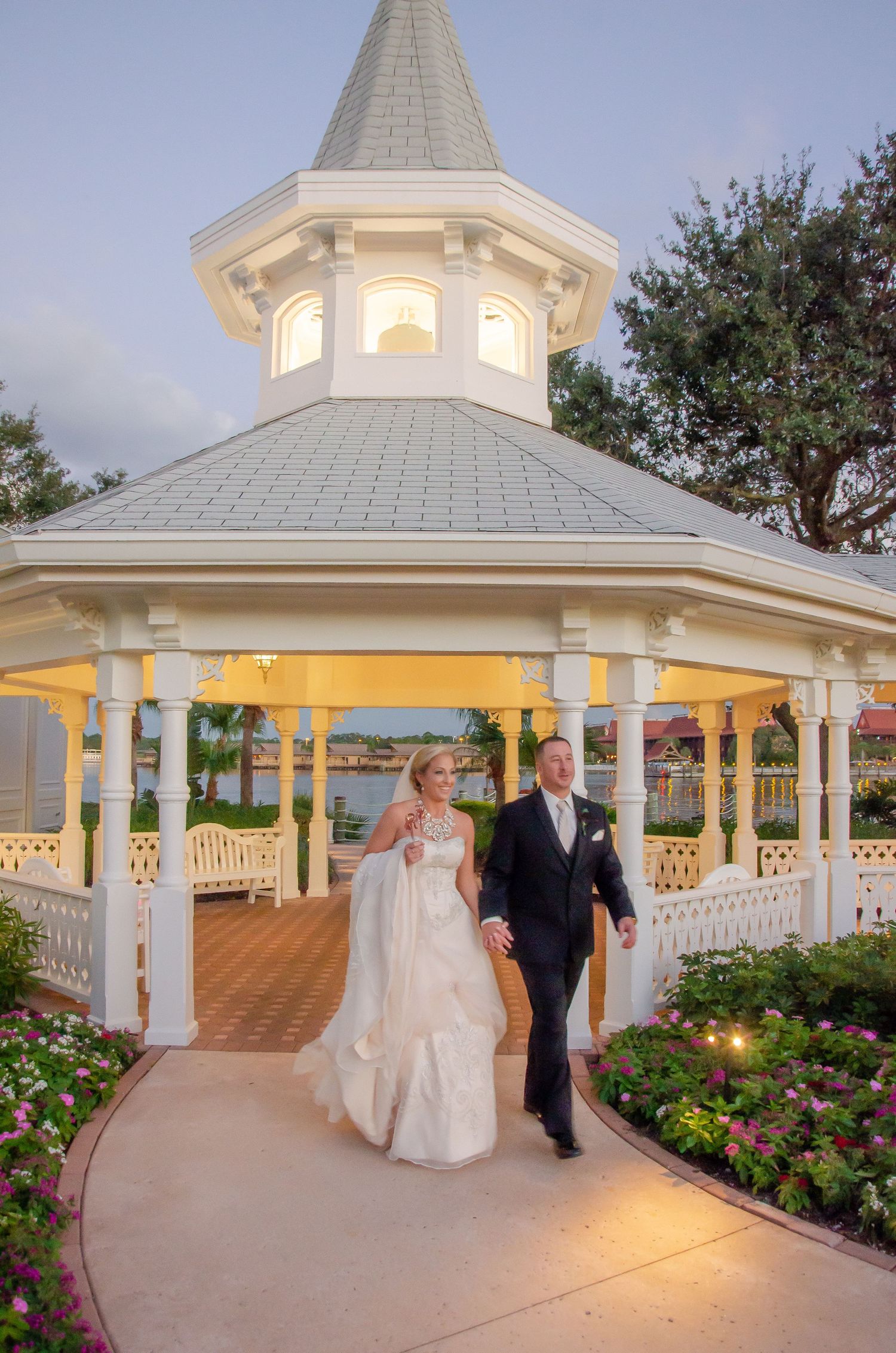 Bride and groom walk hand-in-hand from a elegant white gazebo at dusk during their wedding celebration.