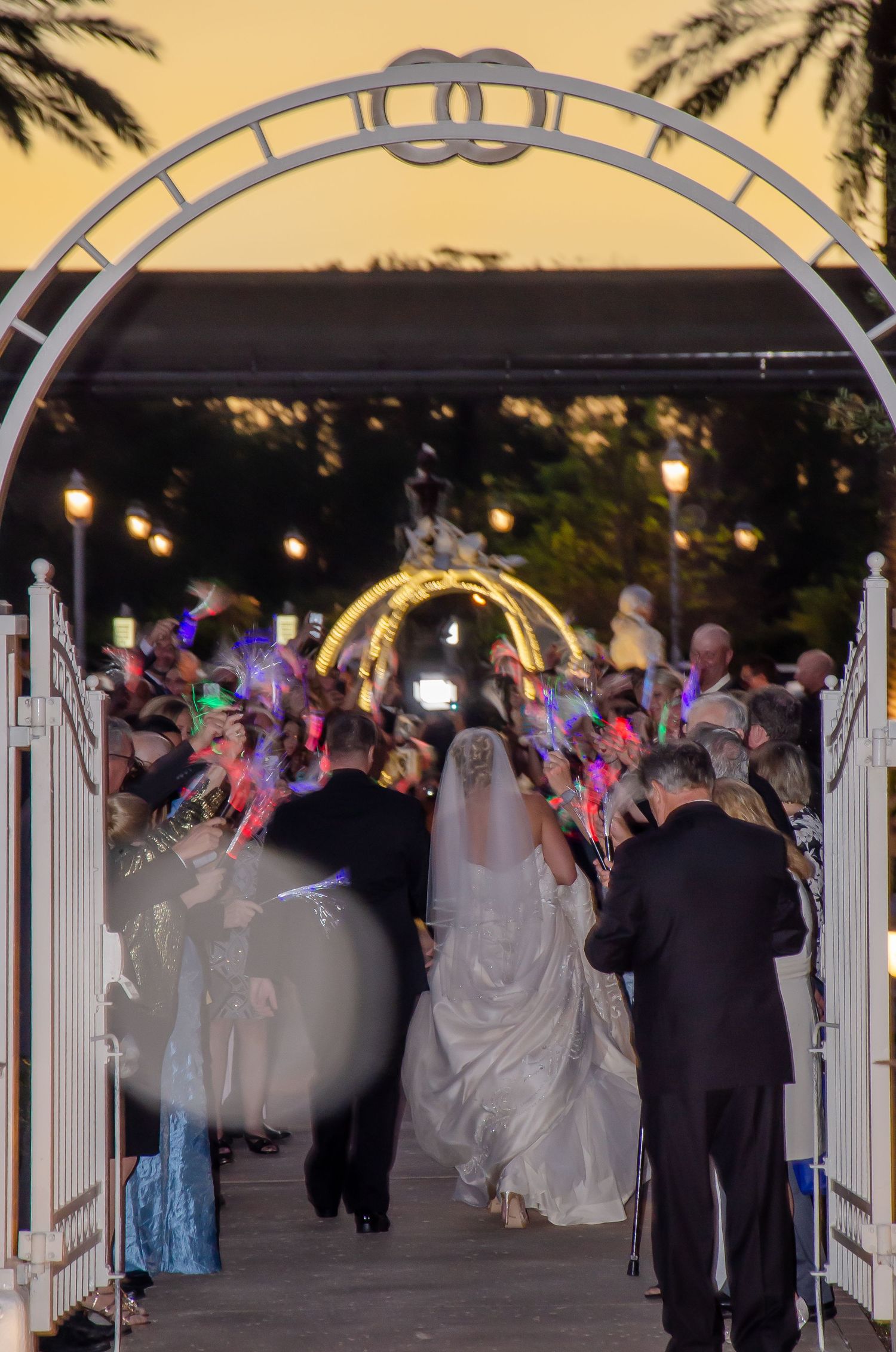 Bride and groom exit wedding ceremony under white arch with glowing crowd at sunset.
