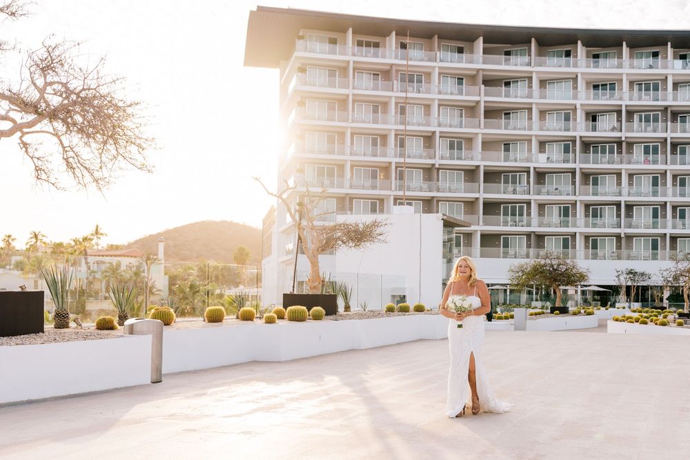 Bride in white gown holding bouquet stands in front of a modern white hotel with cacti at golden hour.