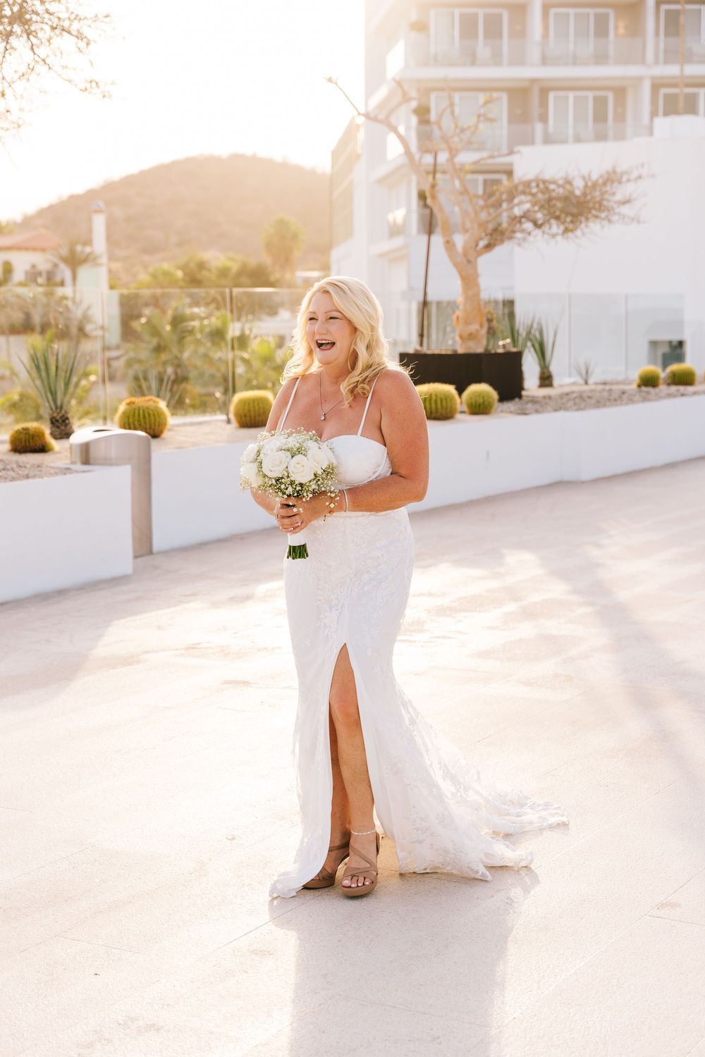 Smiling bride in white slit gown holds bouquet outdoors at golden hour near desert resort.