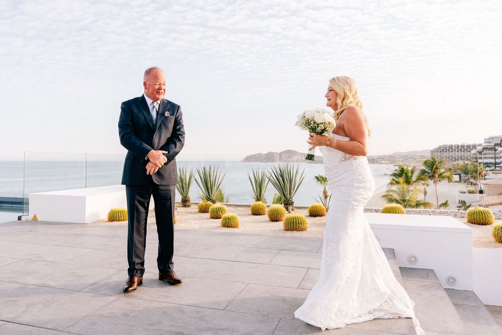 Bride in white gown holds bouquet as groom in navy suit reacts during first look at oceanside wedding venue.