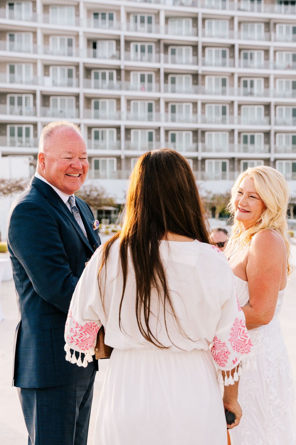 A smiling couple exchanges vows with an officiant in a white dress outside a beachfront hotel.